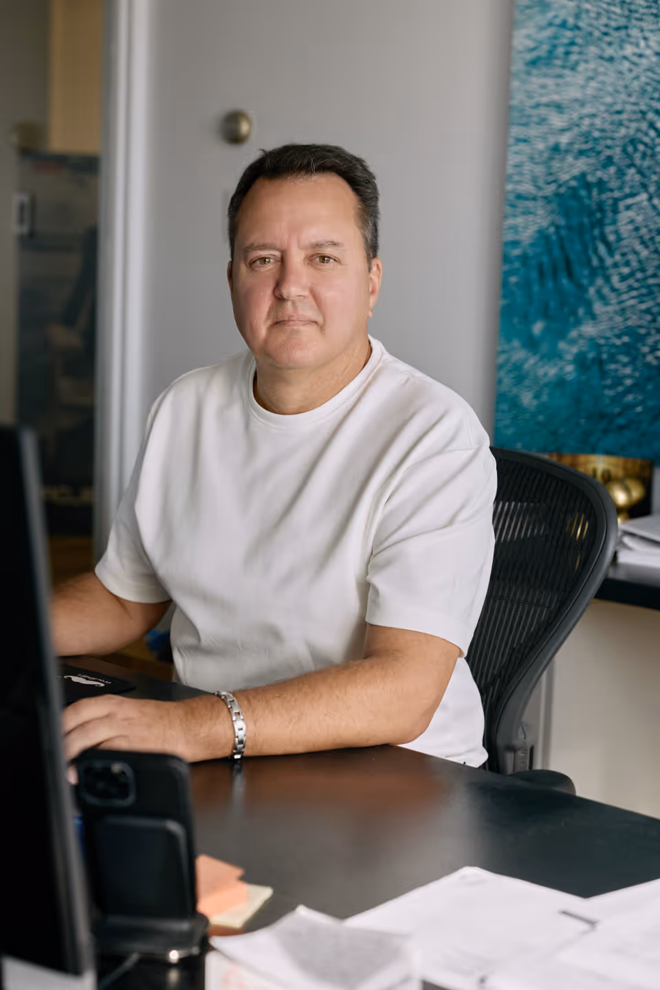 Man in a white t-shirt sitting at a desk with papers and a smartphone, looking at the camera.