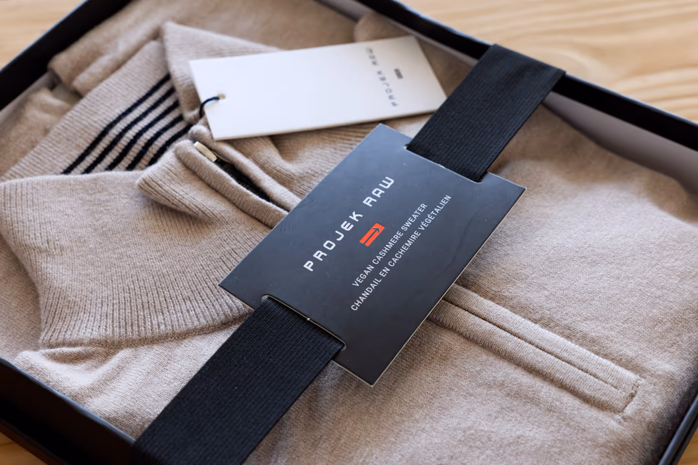 Beige vegan cashmere sweater folded in a box with a black Projek Raw label and a white tag.