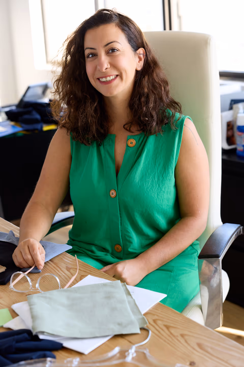 Smiling woman with curly hair wearing a green sleeveless top sitting at a desk with fabric samples and eyeglasses.