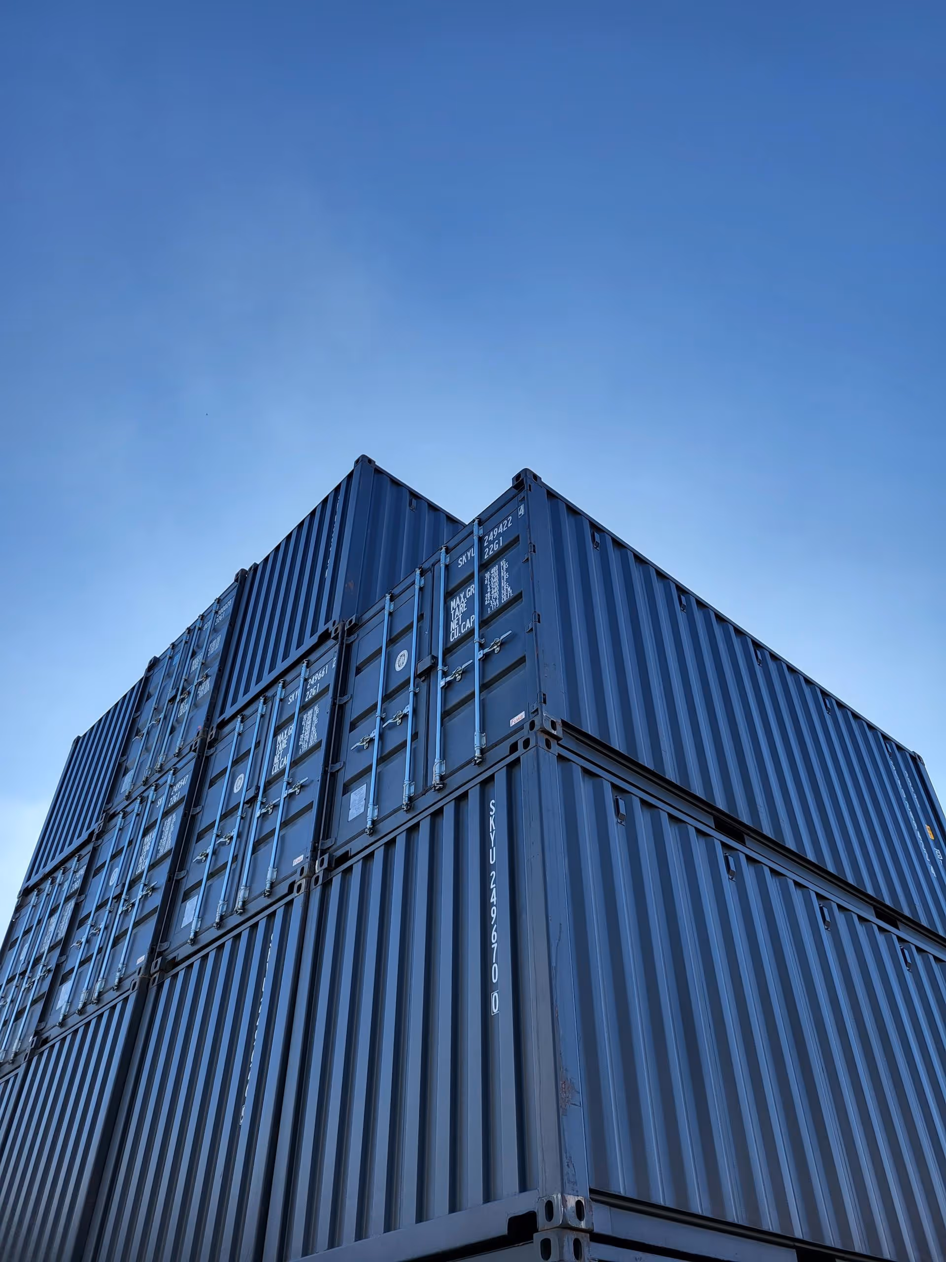 Stacked blue shipping containers viewed from below against a clear blue sky.