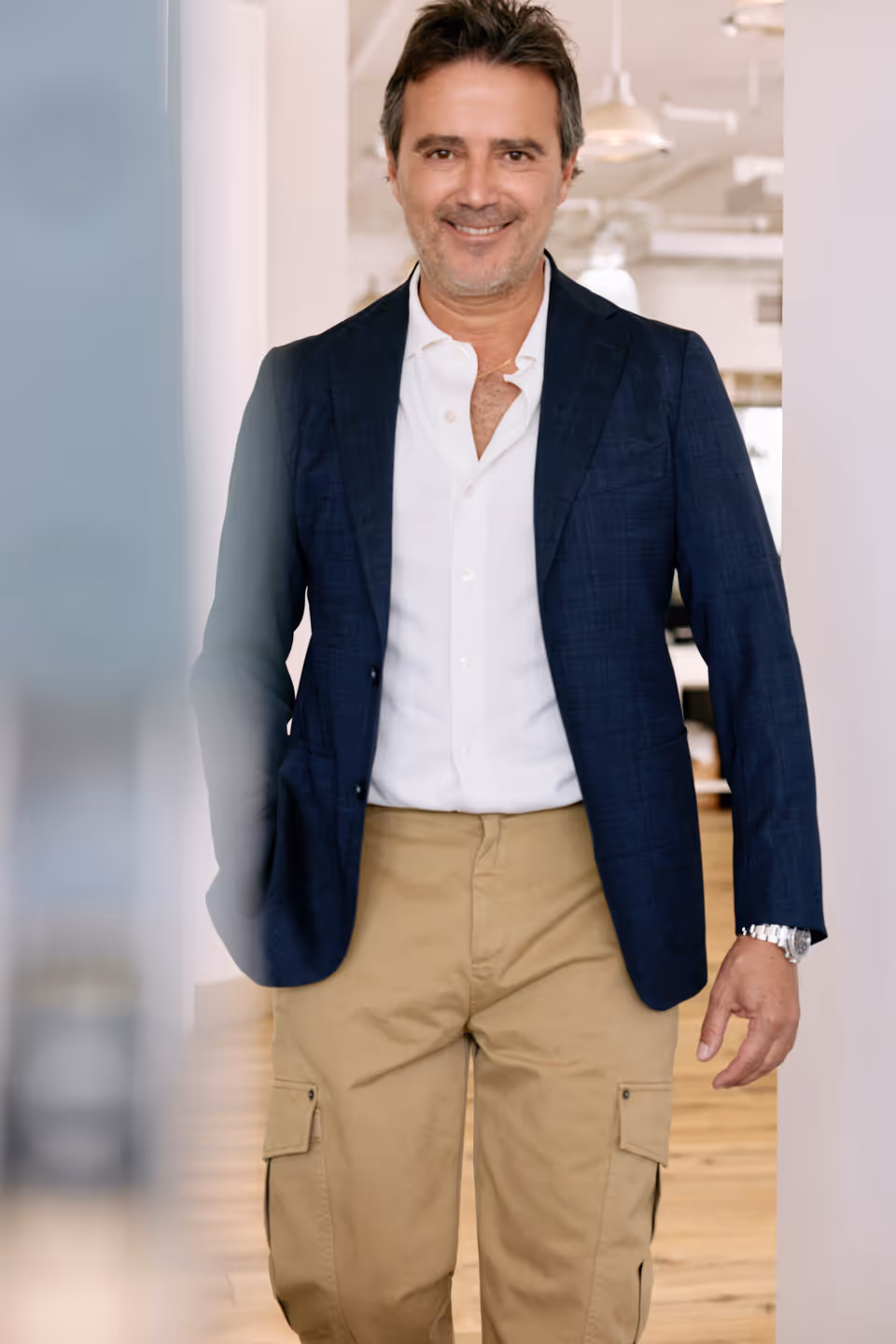 Smiling man wearing a navy blazer, white shirt, and beige cargo pants standing indoors.