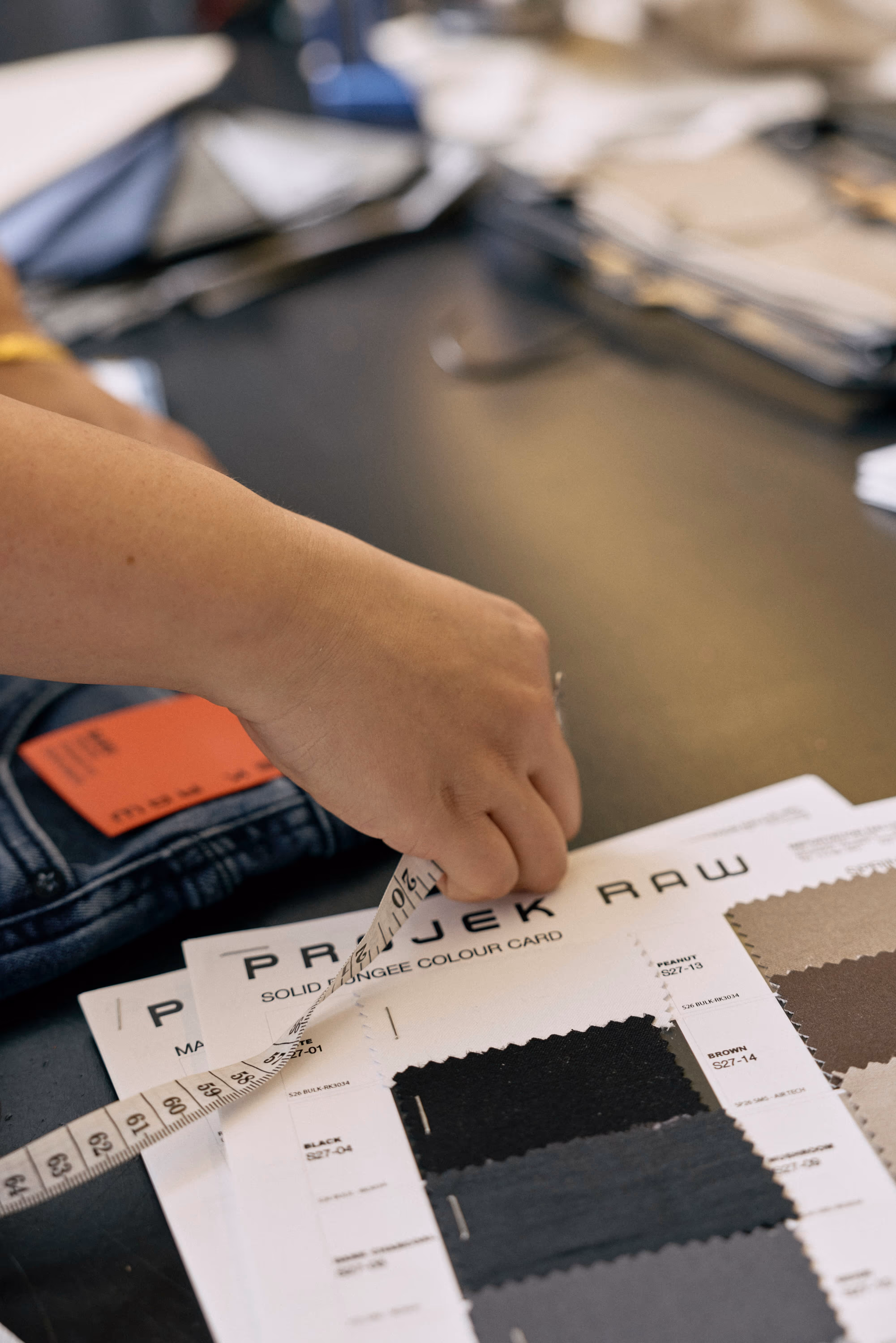 Hand holding a measuring tape over fabric color swatches labeled 'PROJEK RAW' on a table.