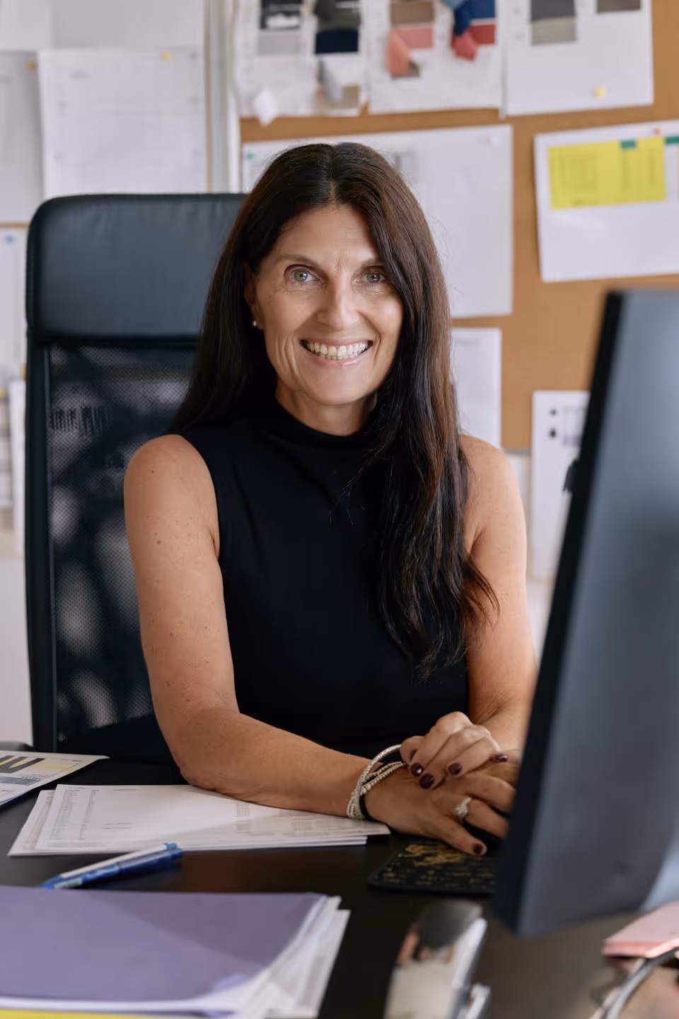 Smiling woman with long dark hair sitting at a desk in an office, hands clasped and papers spread out.