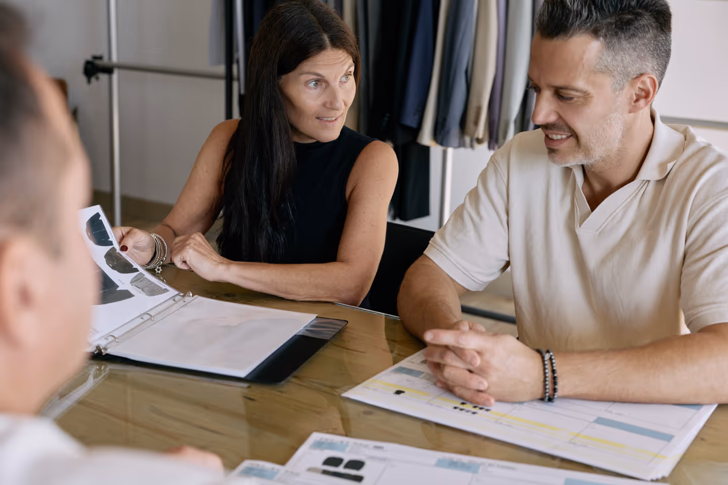 A woman and a man sitting at a table reviewing documents with clothing samples, discussing with a third person.