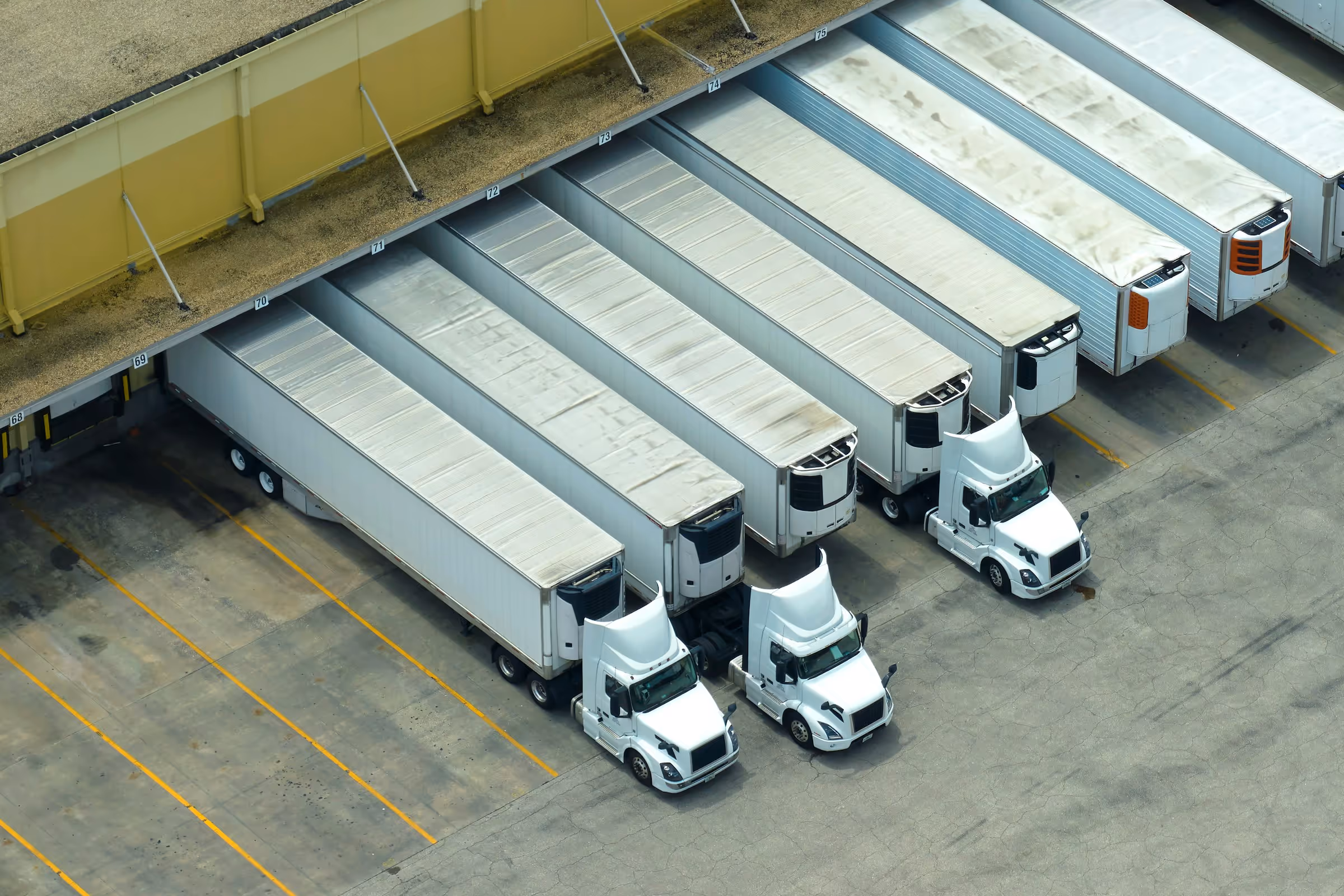 Aerial view of six white semi-trailer trucks parked in loading bays at a warehouse.