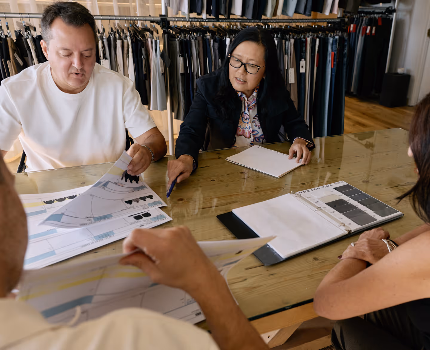 Four people sitting around a wooden table reviewing clothing design sketches and fabric samples, with racks of pants in the background.