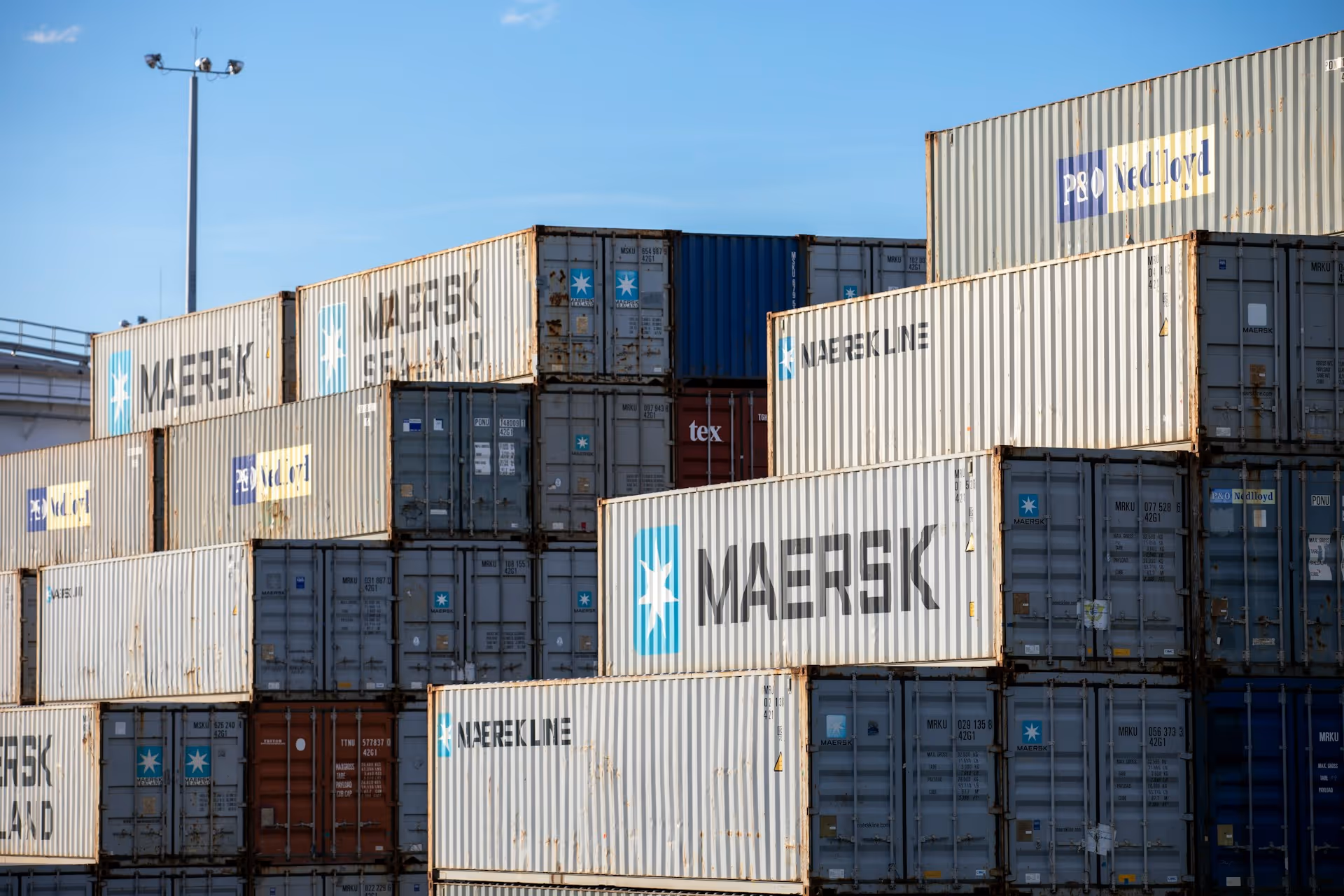 Stacked shipping containers with Maersk, P&O Nedlloyd, and Naersekline logos under a clear blue sky.
