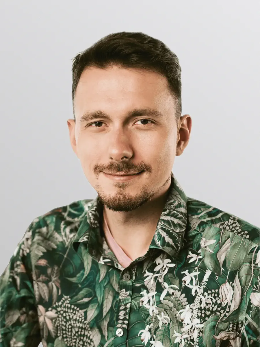 Smiling man with short dark hair and light facial hair wearing a green floral patterned shirt against a plain light background.