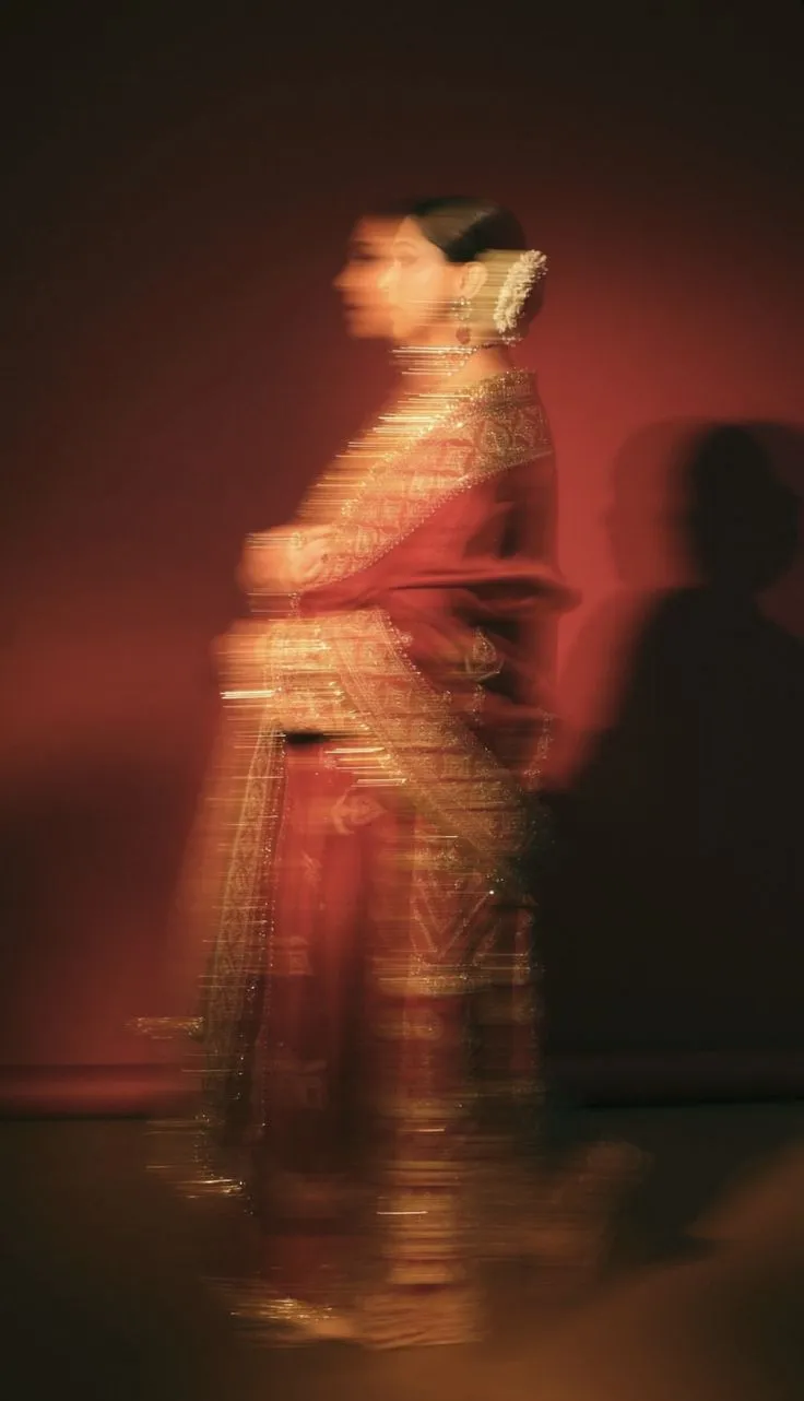 Blurred side profile of a woman wearing a traditional red and gold embroidered sari with floral hair accessories against a dark red background.