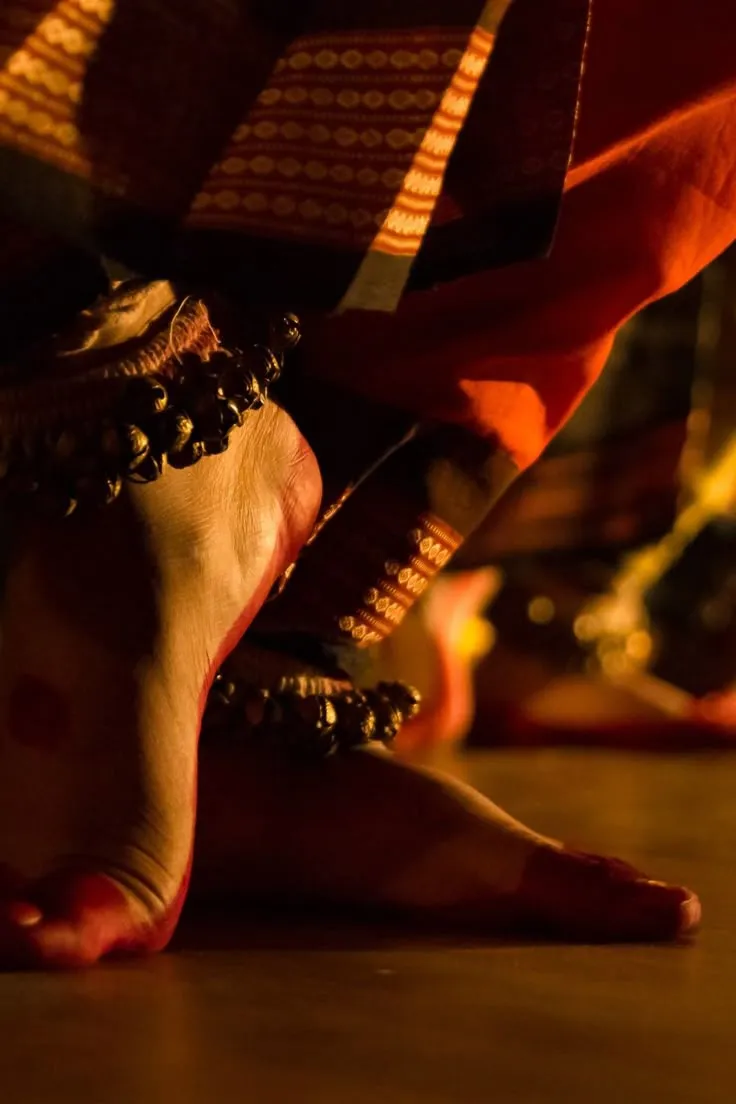 Close-up of bare feet adorned with traditional ankle bells and red attire, poised on a floor during a classical dance performance.