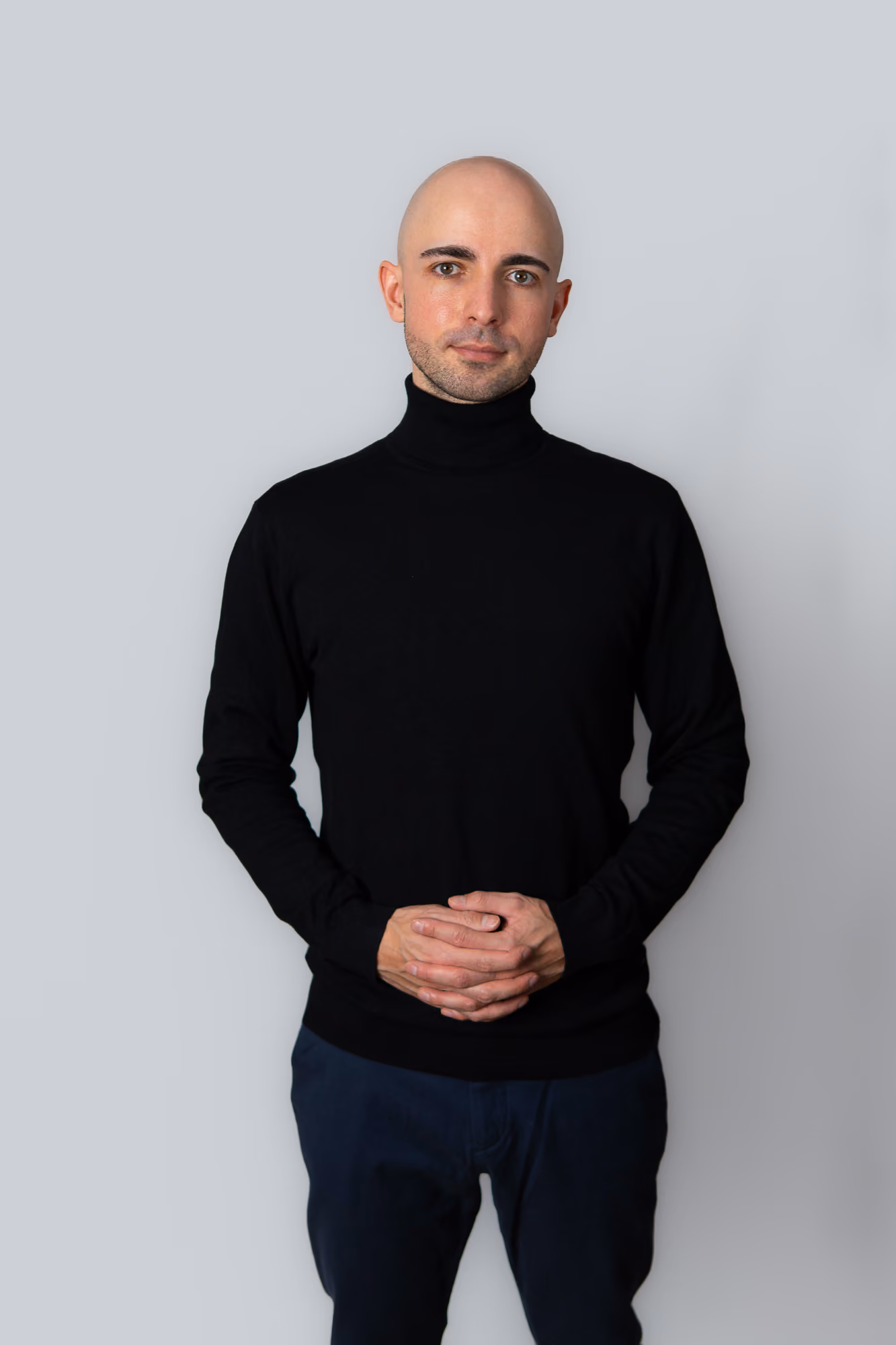 Ein Mann mit Glatze, schwarzem Rollkragenpullover und blauen Hosen vor grauem Hintergrund.