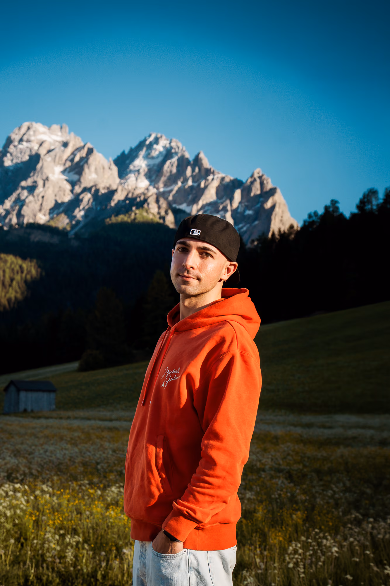 Junger Mann mit rotem Hoodie und schwarzer Kappe vor Berglandschaft mit blauem Himmel.