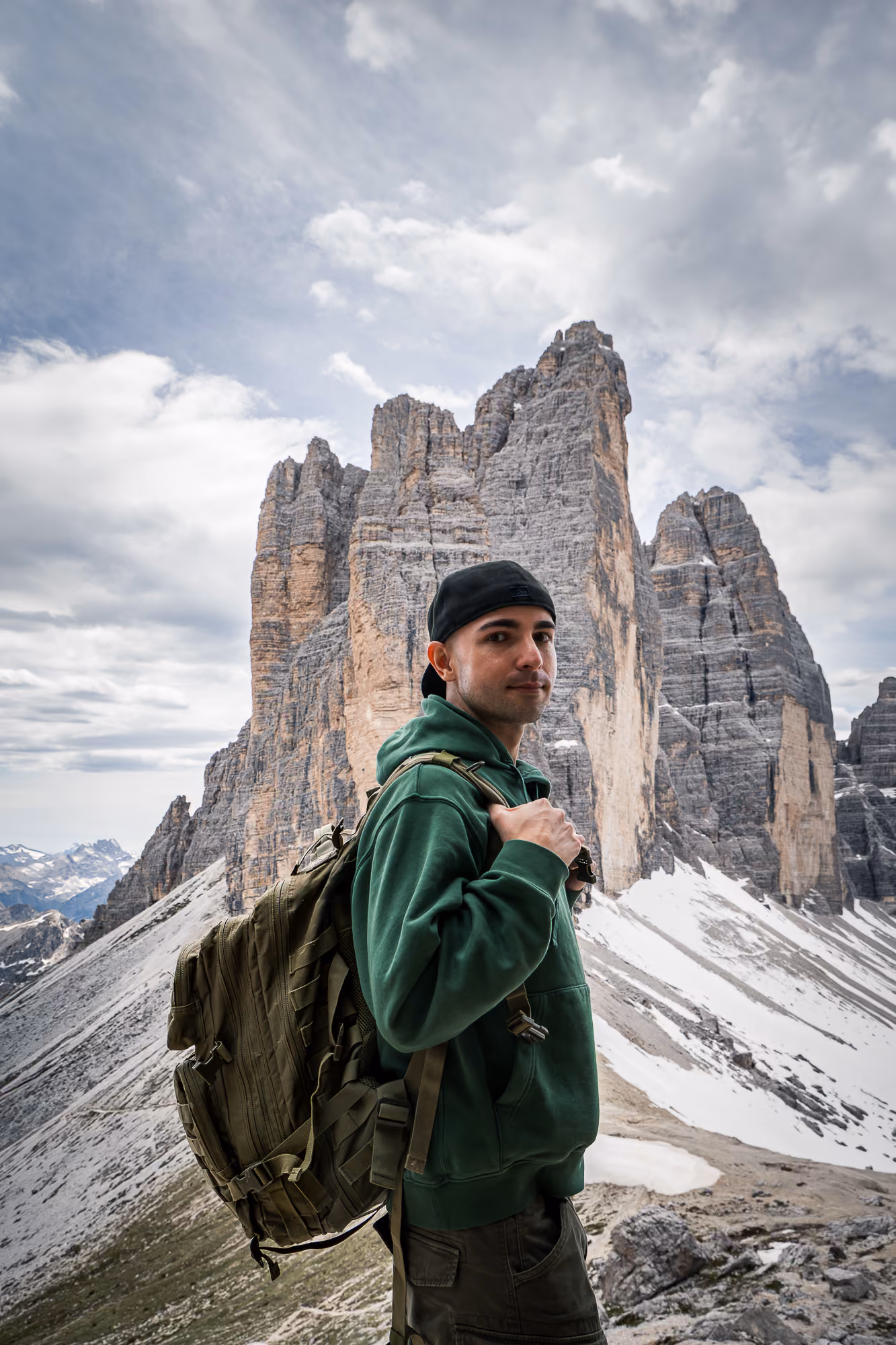 Wanderer mit grüner Kapuzenjacke und Rucksack vor schroffen Berggipfeln mit Schnee.