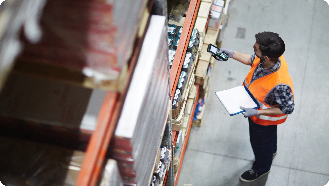 Warehouse worker in an orange safety vest scanning items with a handheld device while holding a clipboard.