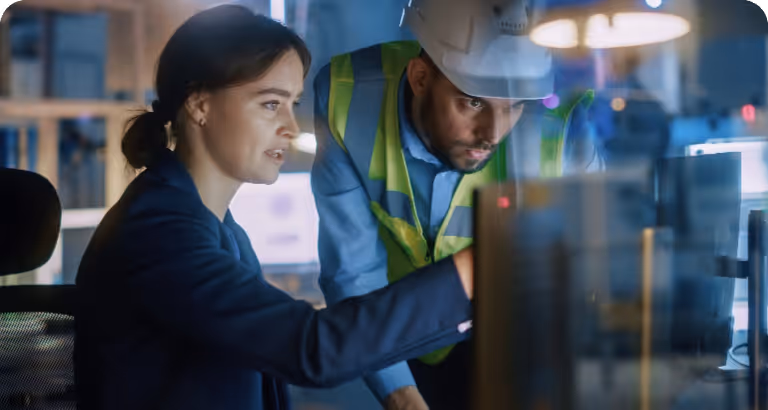 A professional and engineer in a hard hat and safety vest review data on a computer screen in an office.