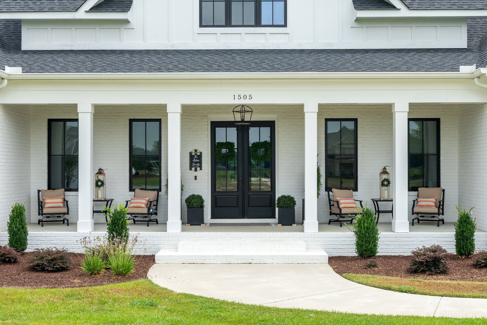 Elegant white farmhouse with black doors, porch chairs, and landscaped entrance