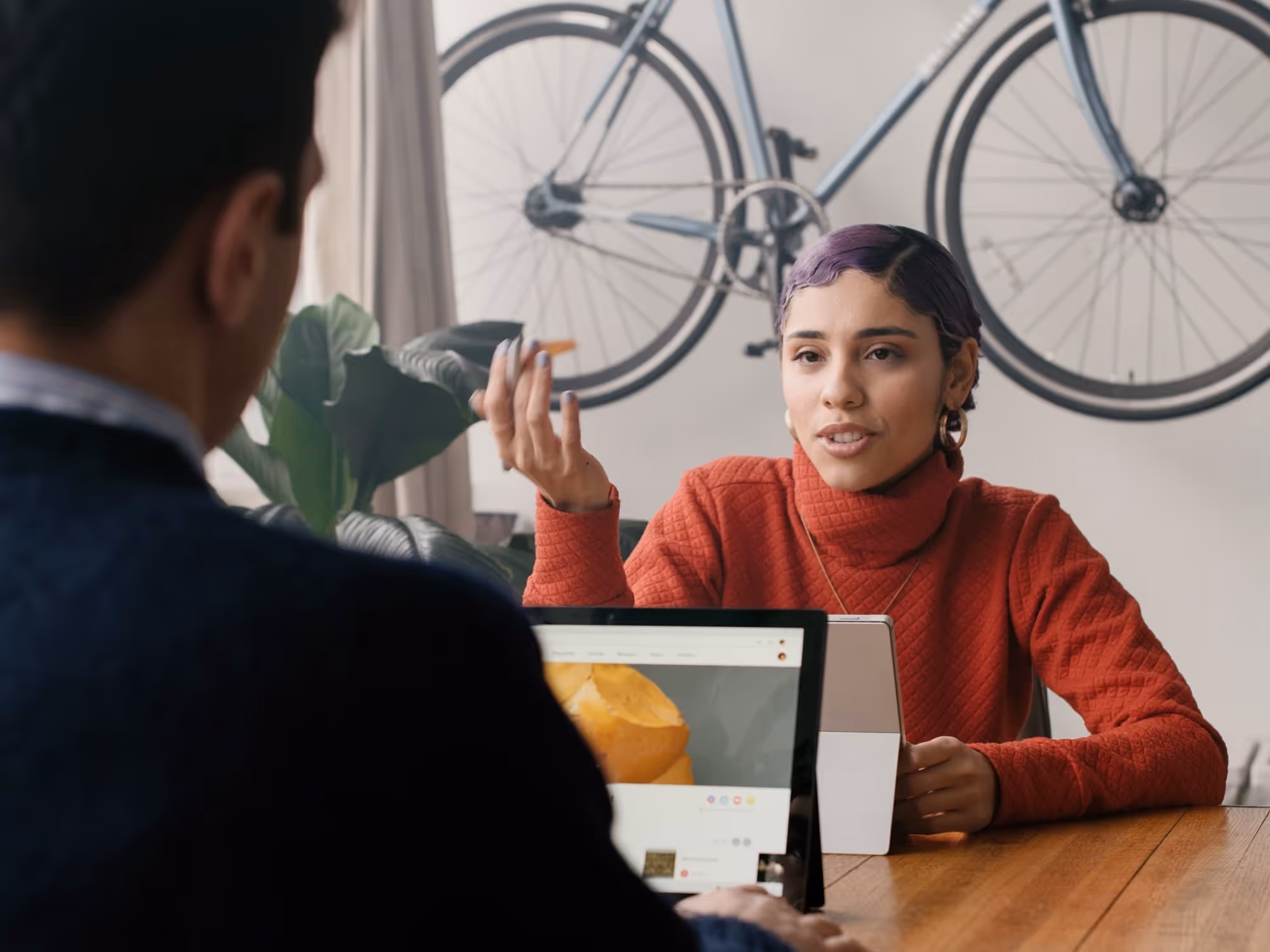 Two people having a conversation at a wooden table, with a bicycle mounted on the wall behind them.