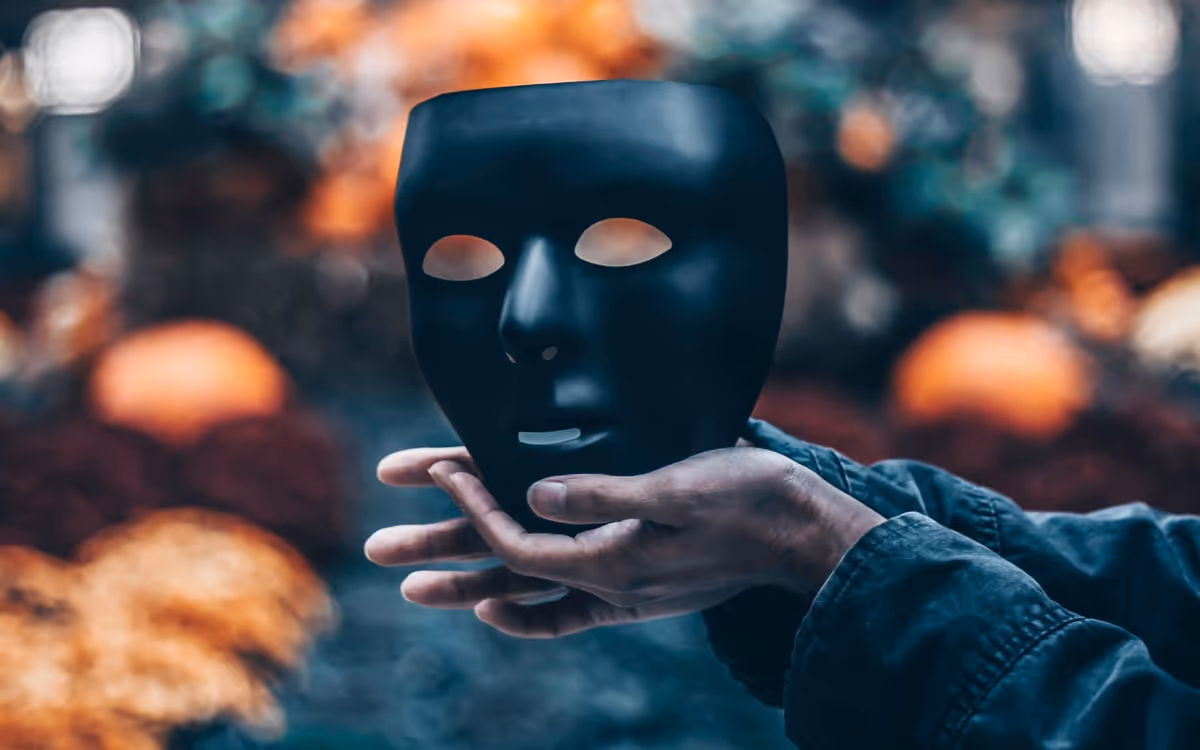 Hands holding a black theatrical mask with eye and mouth cutouts against a blurred background with orange and green hues.