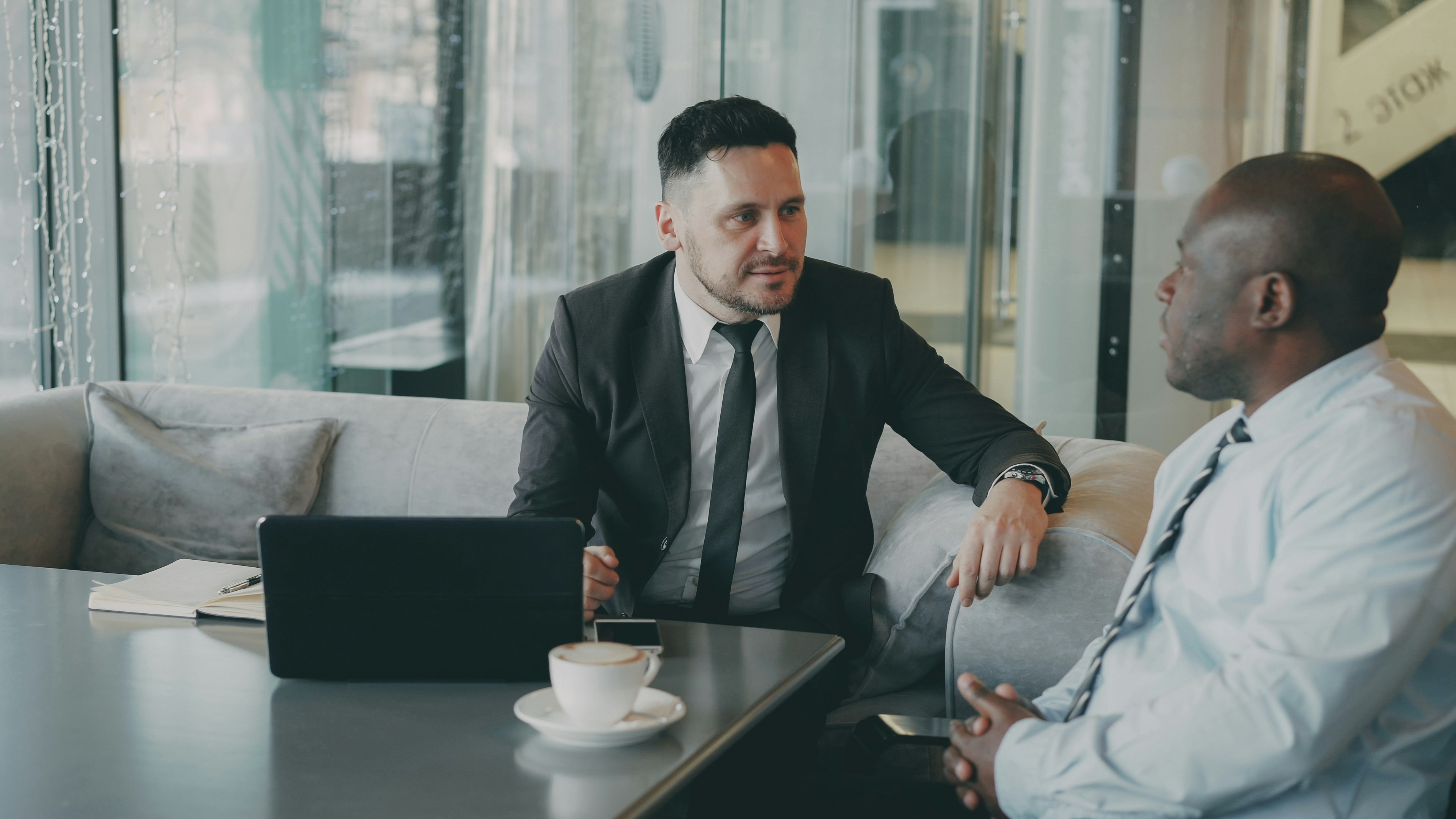 Two businessmen in formal attire having a discussion while seated on a couch with a laptop and coffee on the table.