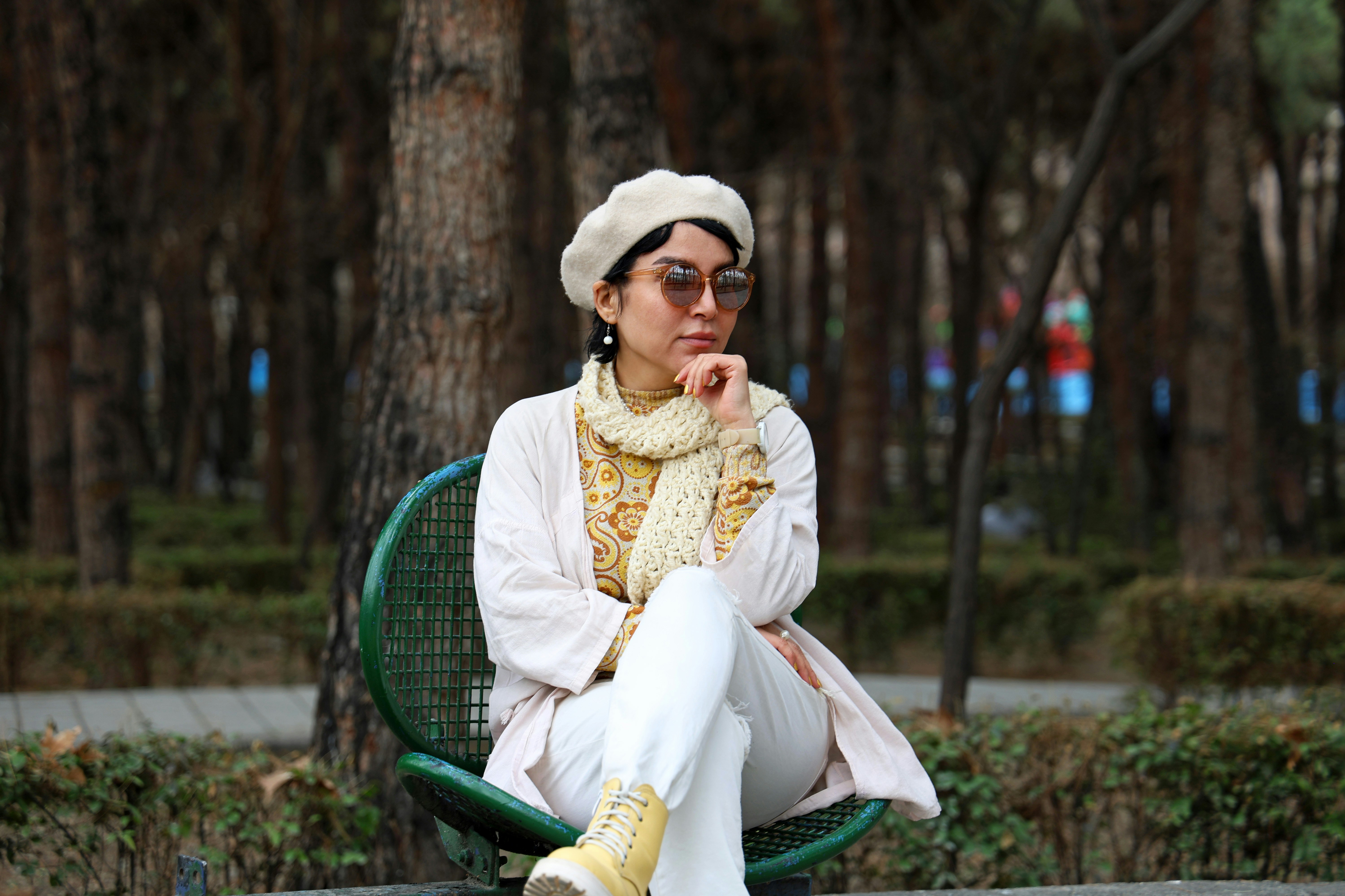A woman wearing a beige beret, patterned yellow top, cream scarf, white pants, and yellow boots sitting on a green chair in a park with trees in the background.