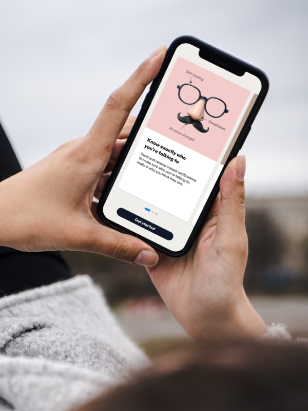 Person holding a smartphone displaying an app screen warning about SIM cloning, deepfakes, and AI voice changers with a message about verifying identity and a 'Get started' button.