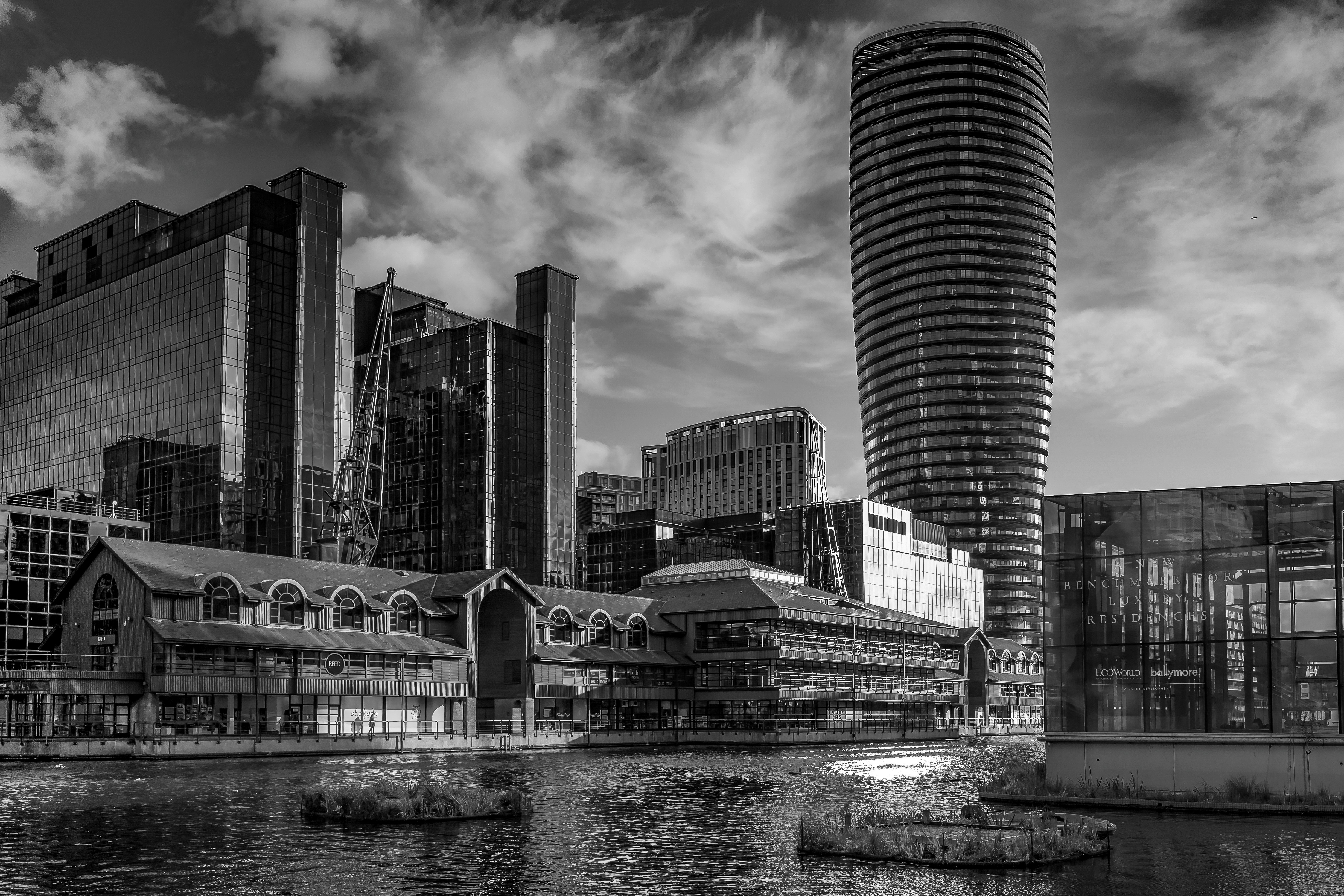 Black and white cityscape featuring modern glass skyscrapers, a round high-rise building, older industrial-style waterside buildings, and a water canal with floating vegetation.