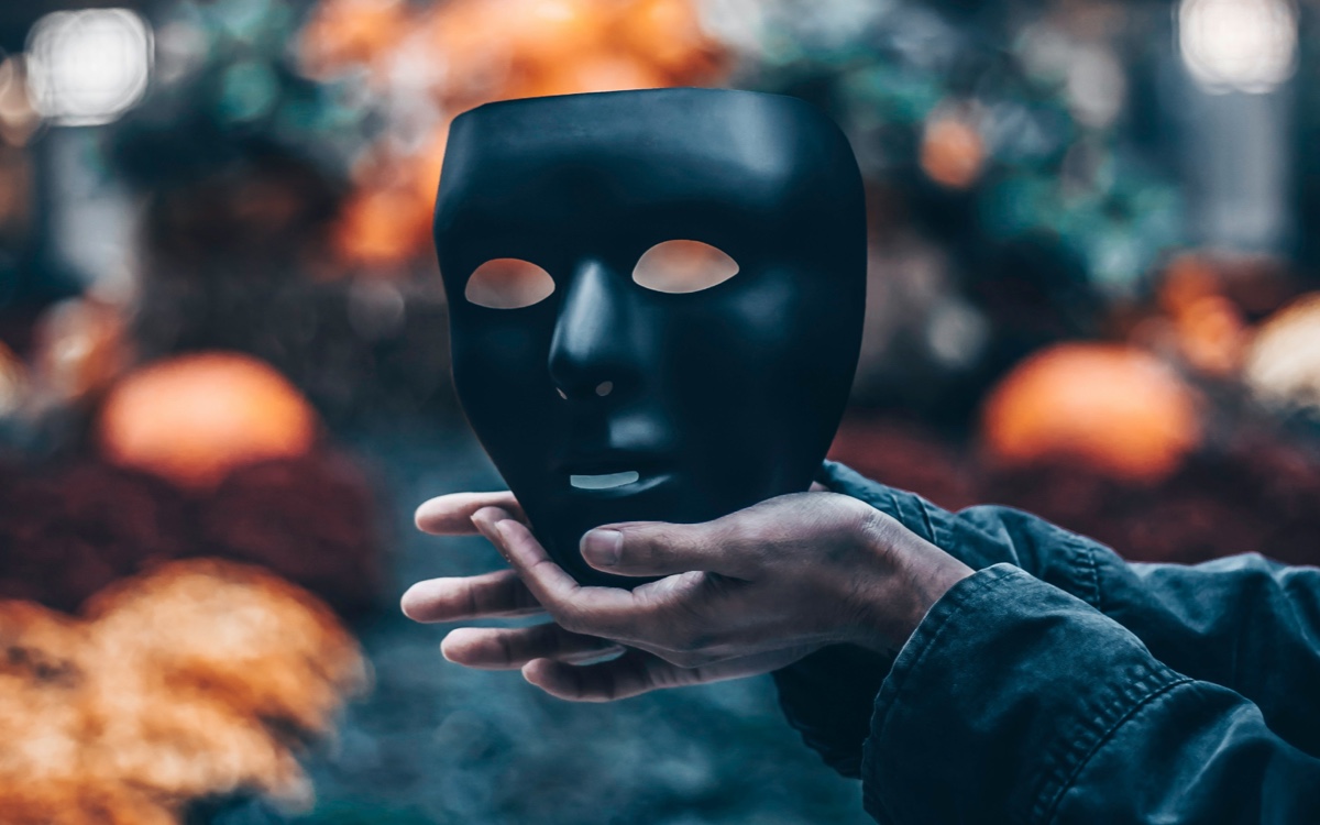 Person holding a black mask with eye and mouth cutouts against a blurred background with warm orange and cool blue tones.