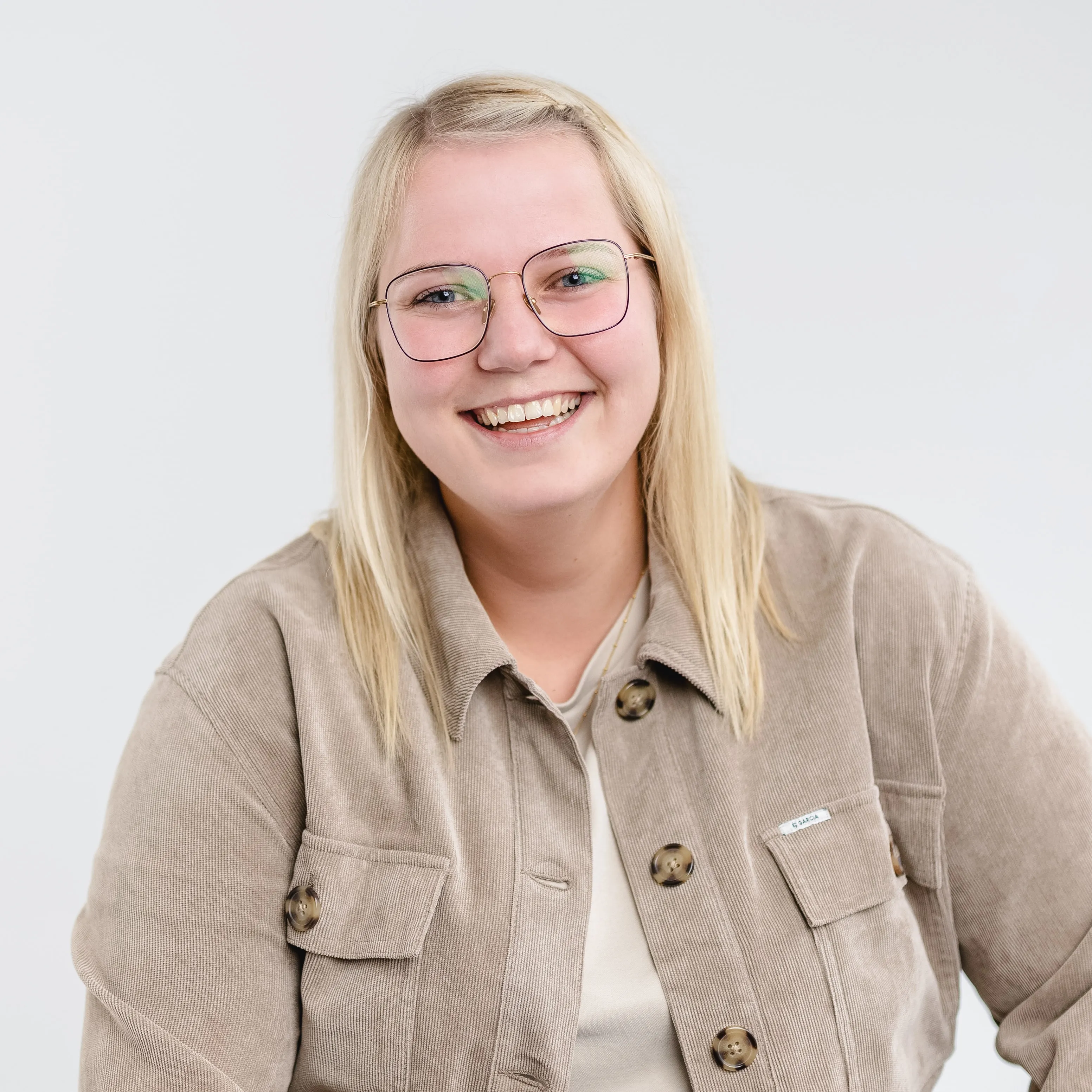 Smiling young woman with blonde hair and glasses wearing a beige jacket against a plain light background.