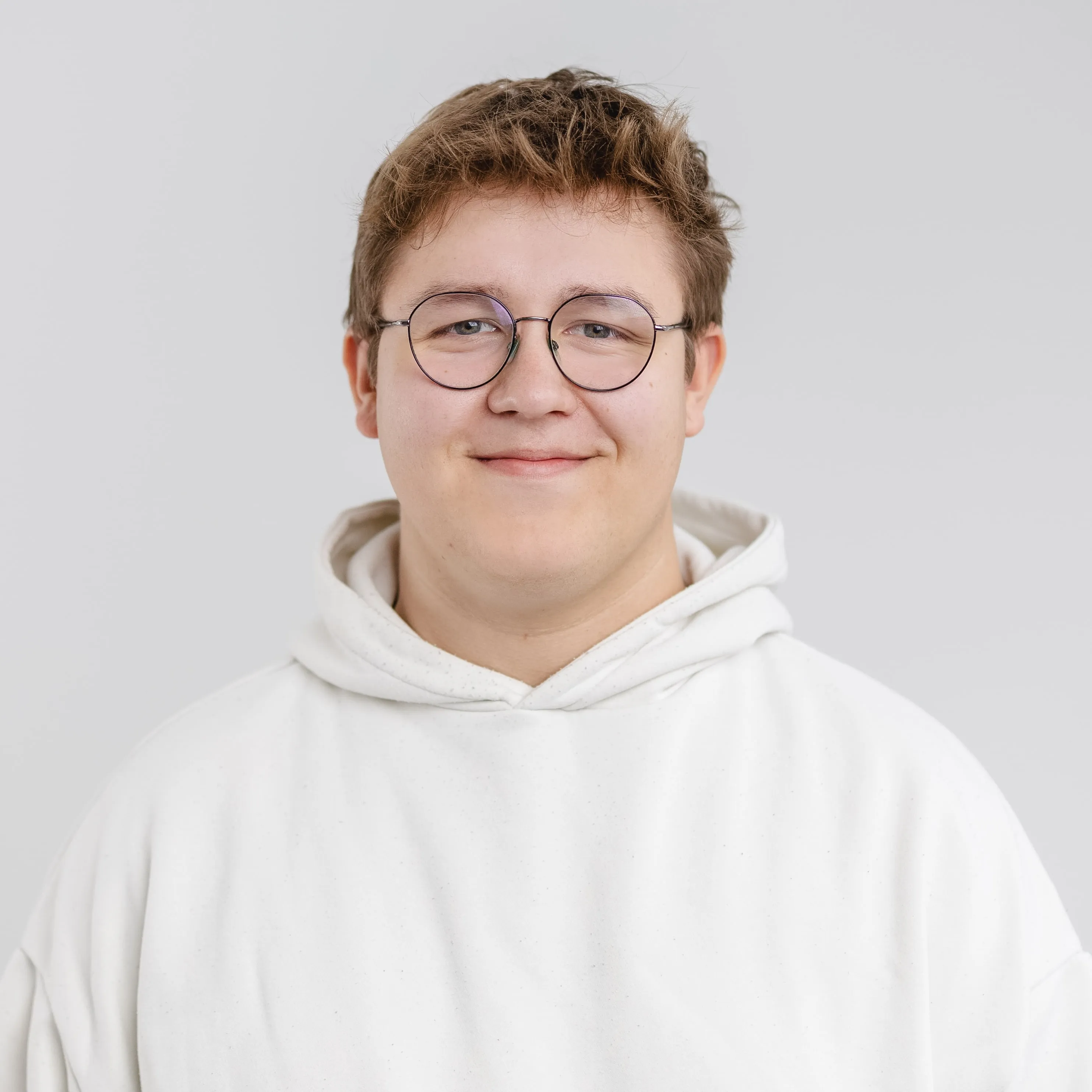 Smiling young man with short brown hair and round glasses wearing a white hoodie against a plain light background.