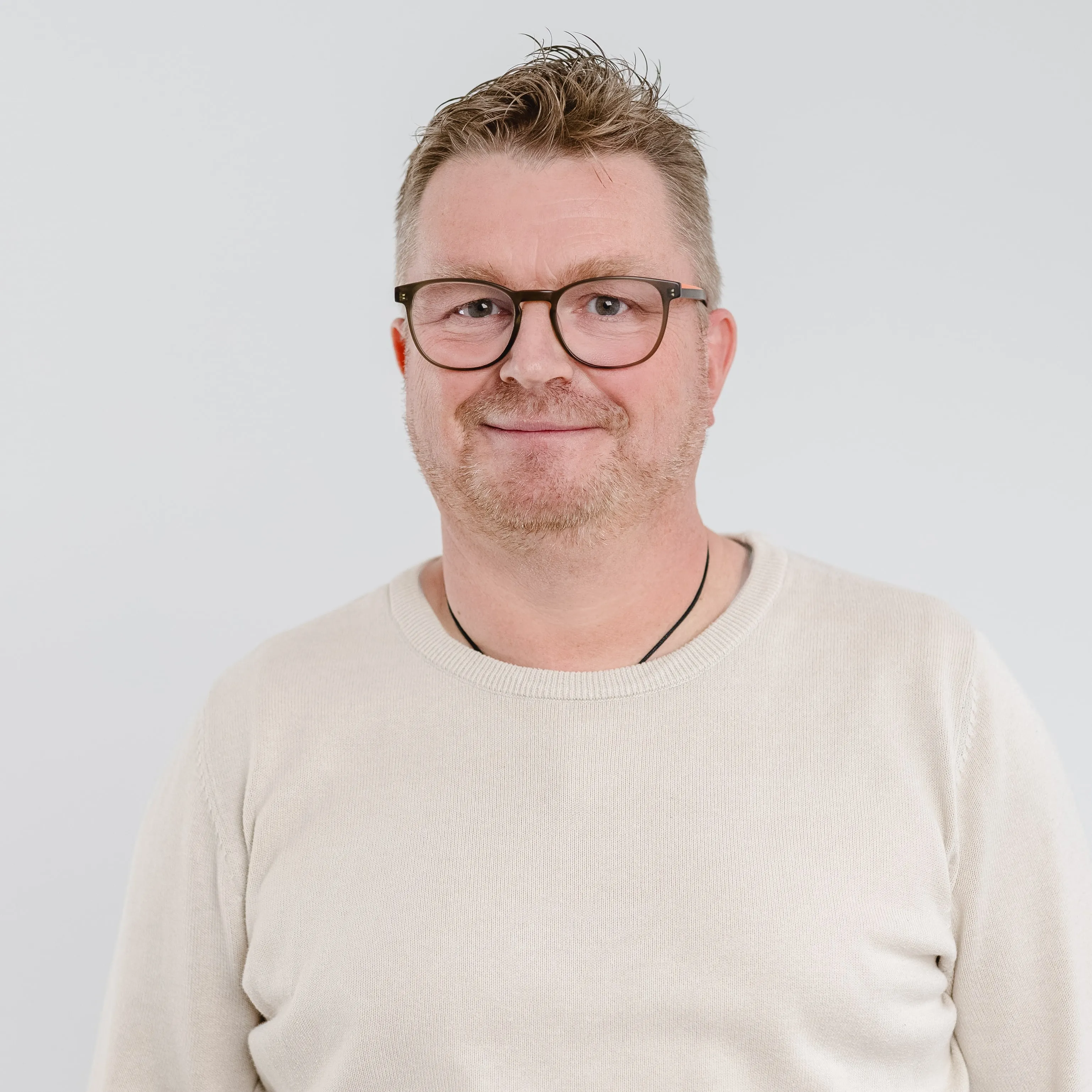 Smiling man with glasses, short blond hair, and light beard wearing a beige sweater and black necklace against a plain white background.