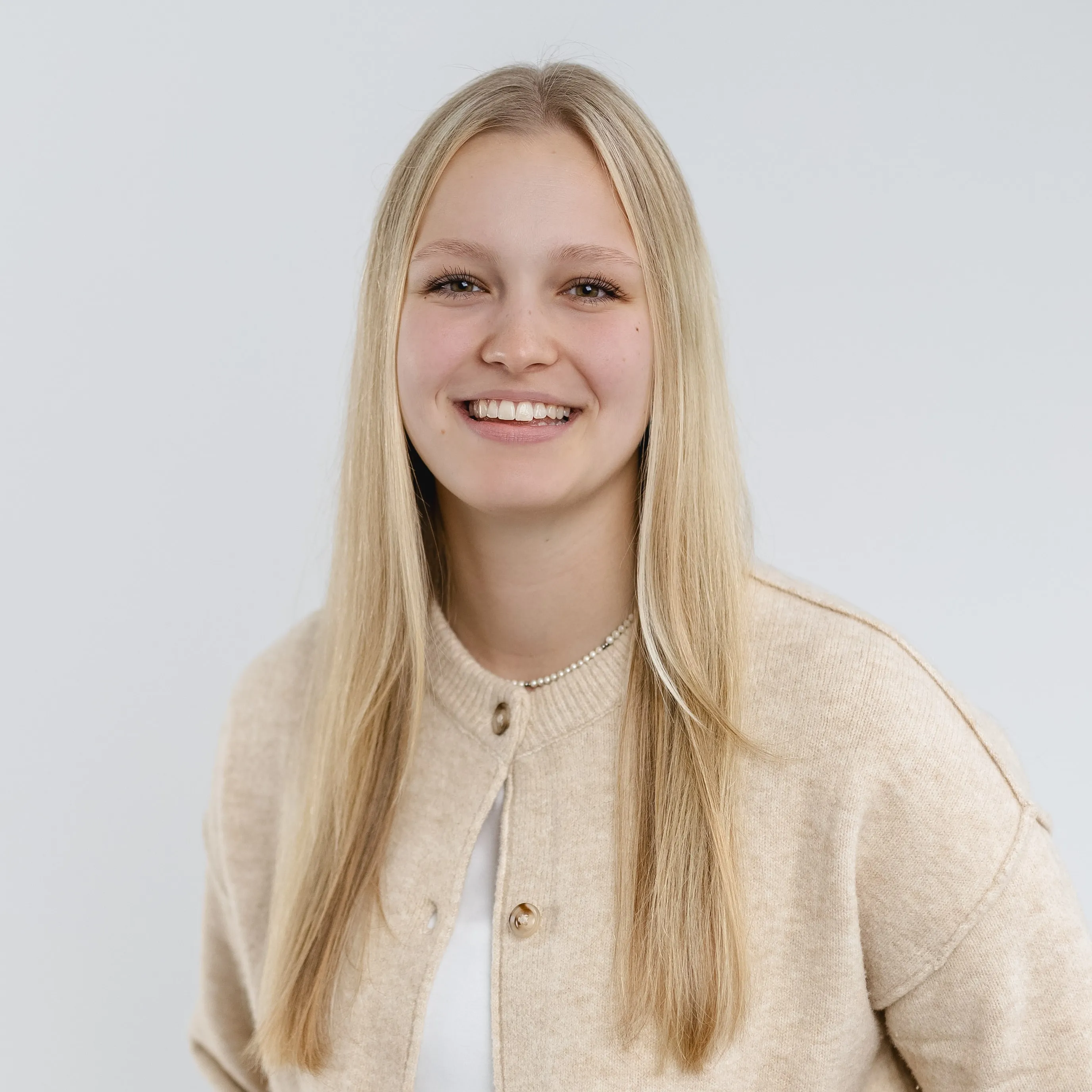 Smiling young woman with long blonde hair wearing a beige cardigan and white shirt against a light background.