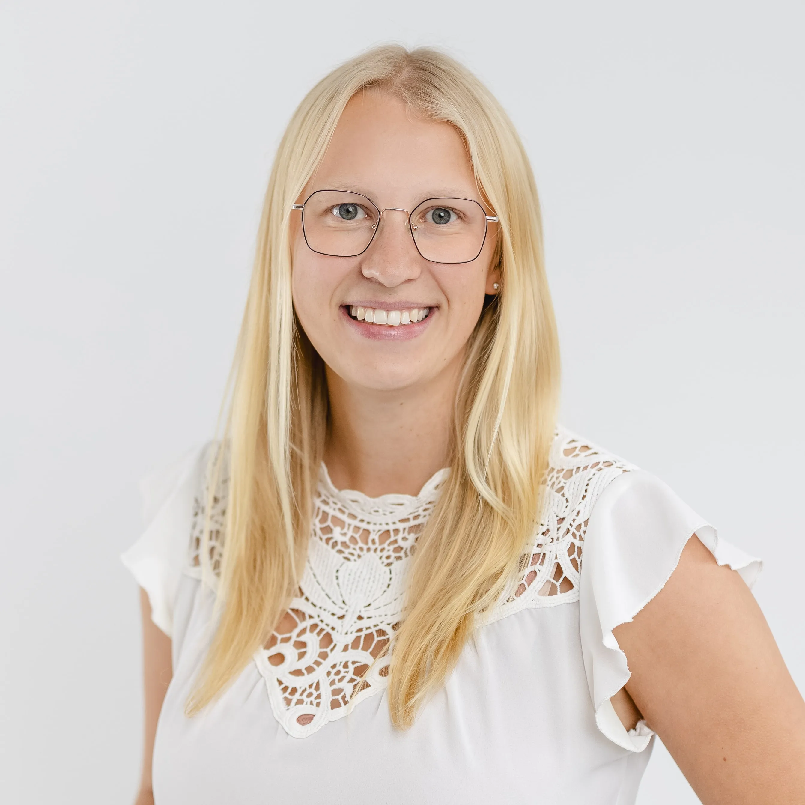 Smiling blonde woman with glasses wearing a white blouse with lace detail against a light background.