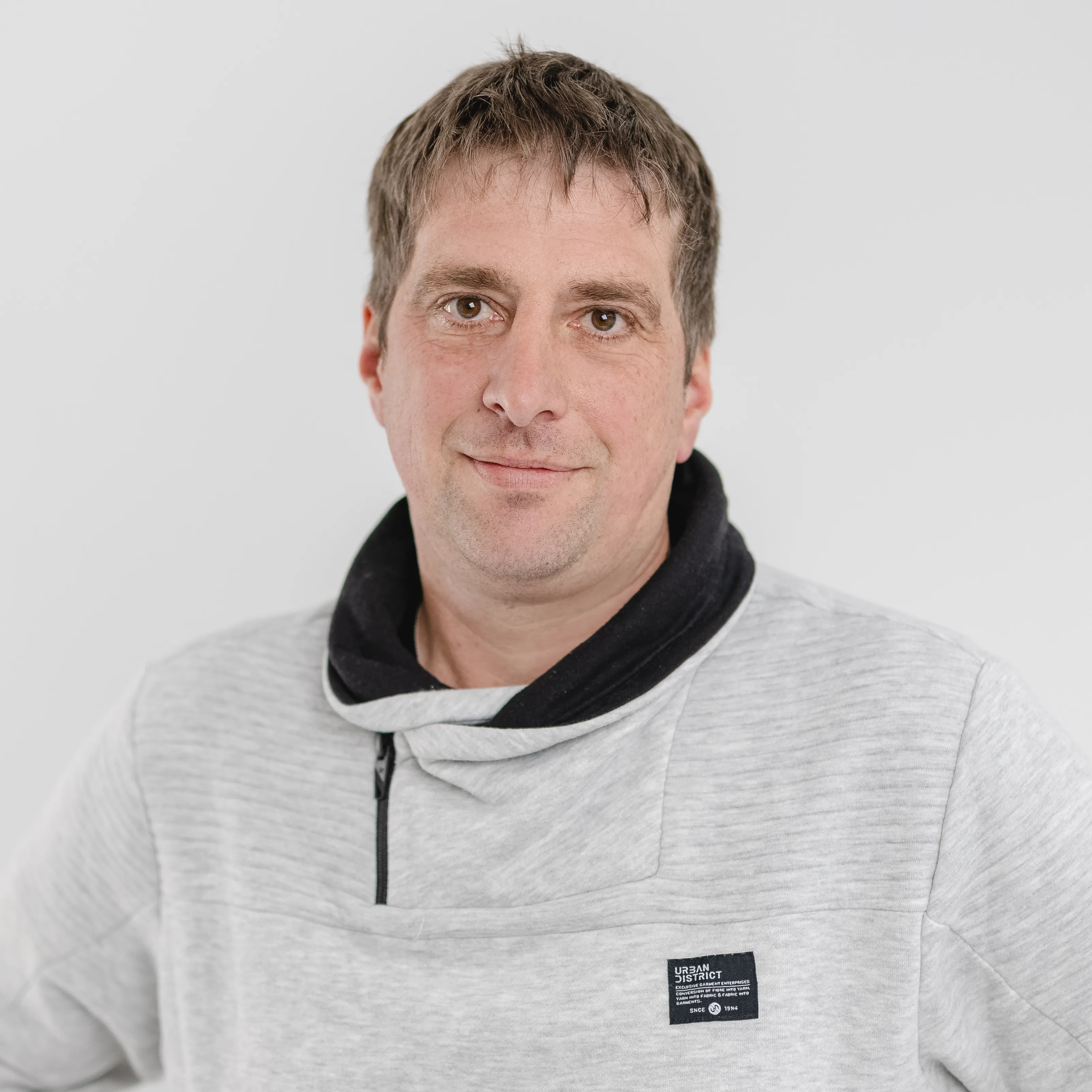 Man with short brown hair wearing a light gray hoodie with a black collar against a plain white background.