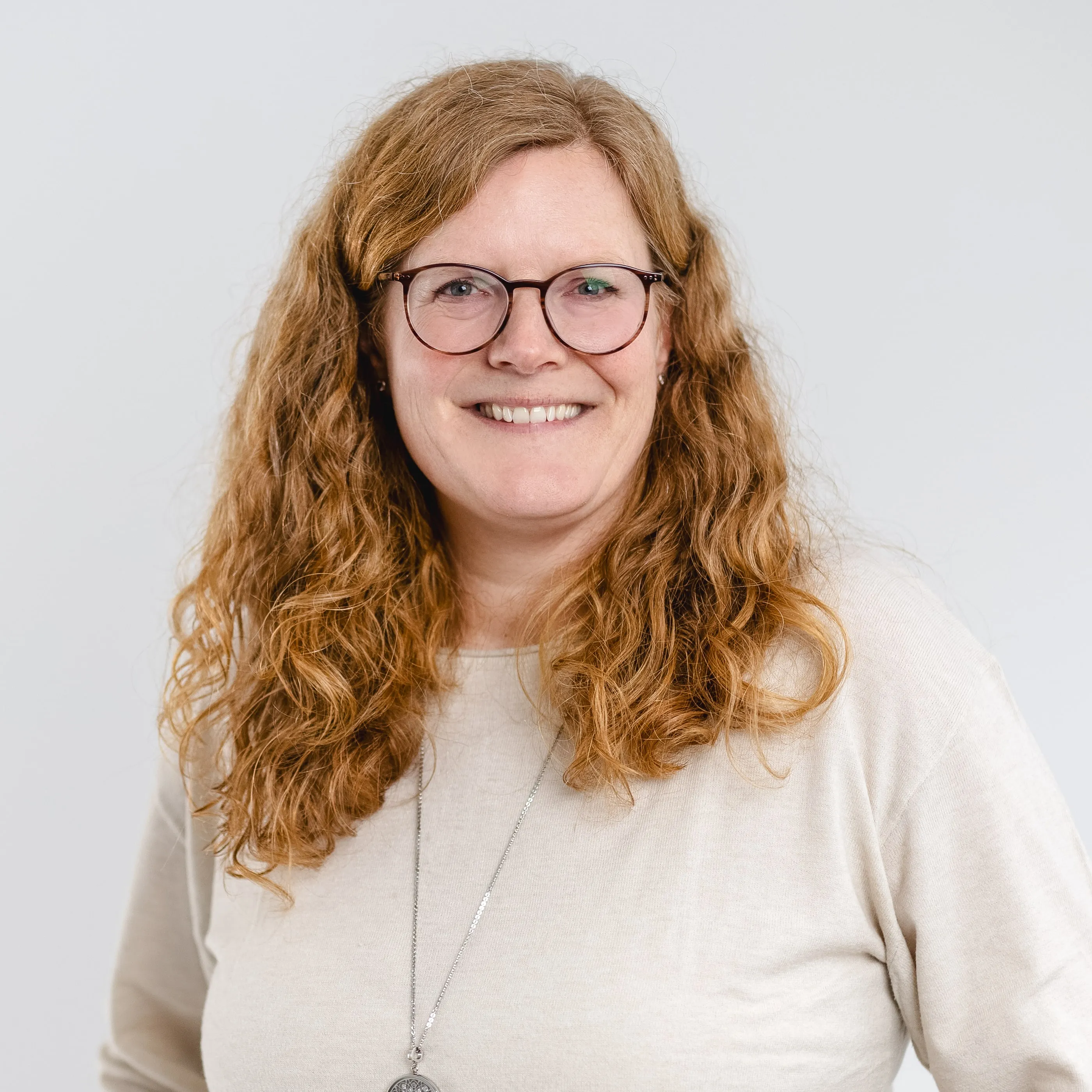 Smiling woman with curly red hair wearing glasses and a beige sweater with a pendant necklace.