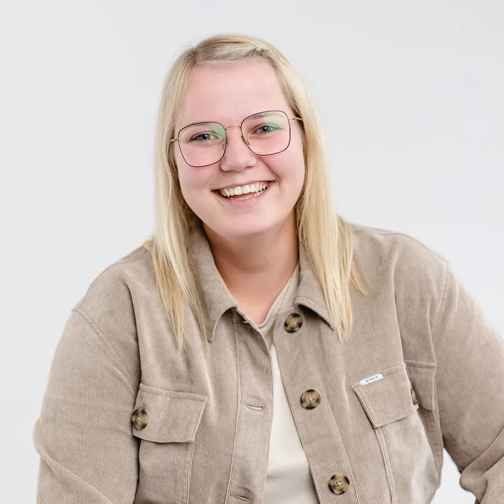 Smiling young woman with blonde hair and glasses wearing a beige jacket against a plain light background.