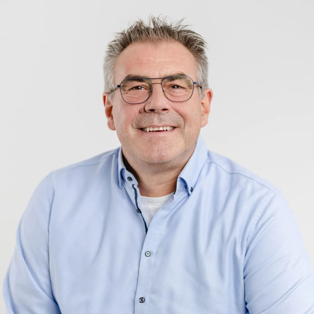 Smiling middle-aged man wearing glasses and a light blue button-up shirt against a plain white background.