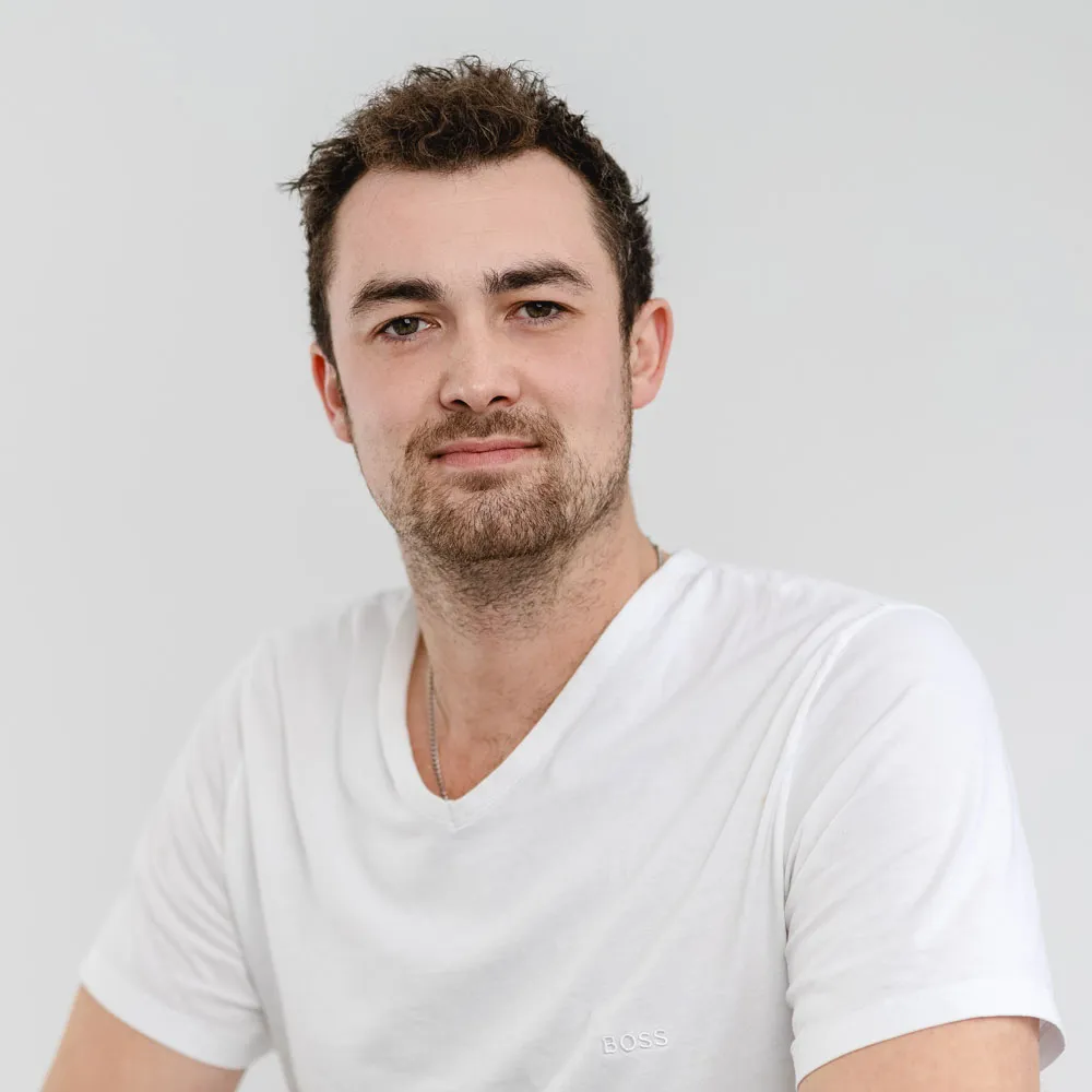 Portrait of a man with short curly hair and light facial hair wearing a white Boss t-shirt against a plain light gray background.