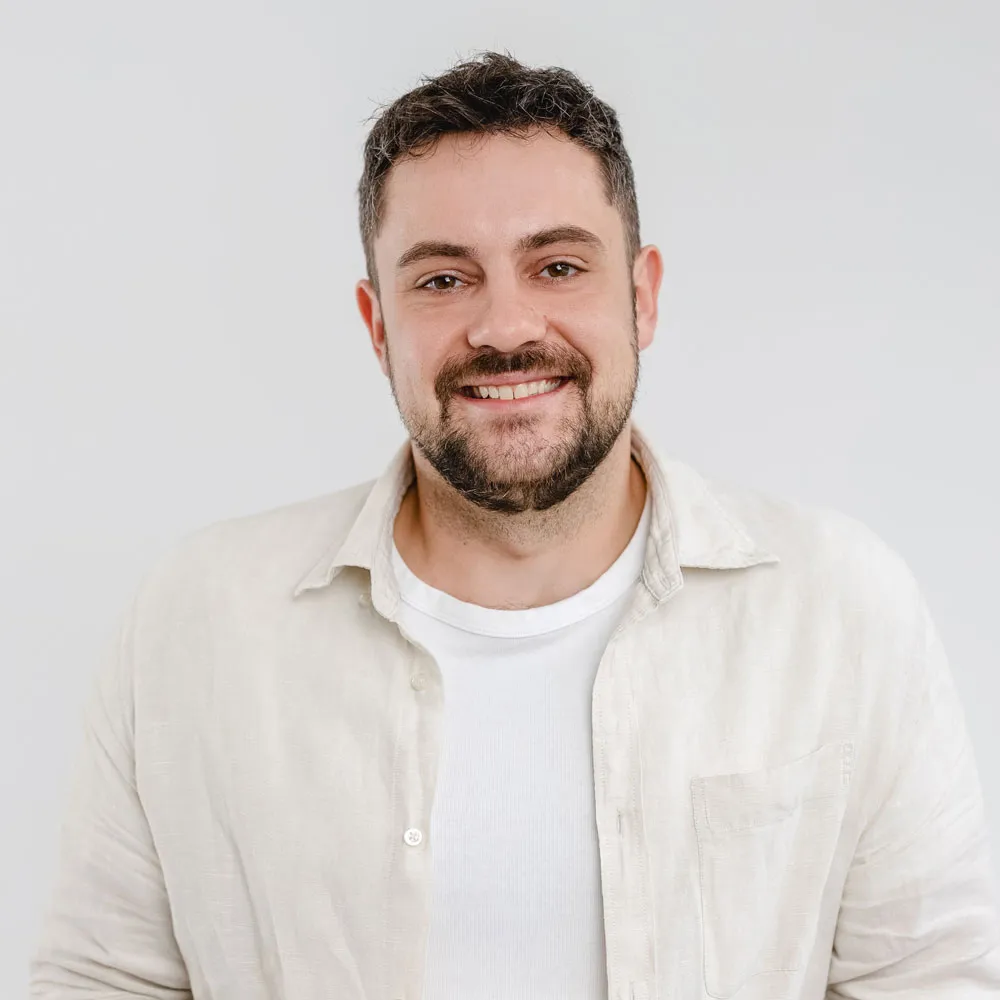 Smiling man with short dark hair and beard wearing a white shirt and beige jacket against a plain background.