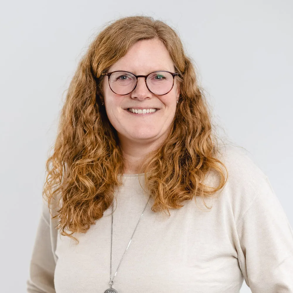Smiling woman with curly red hair wearing glasses and a beige sweater with a pendant necklace.