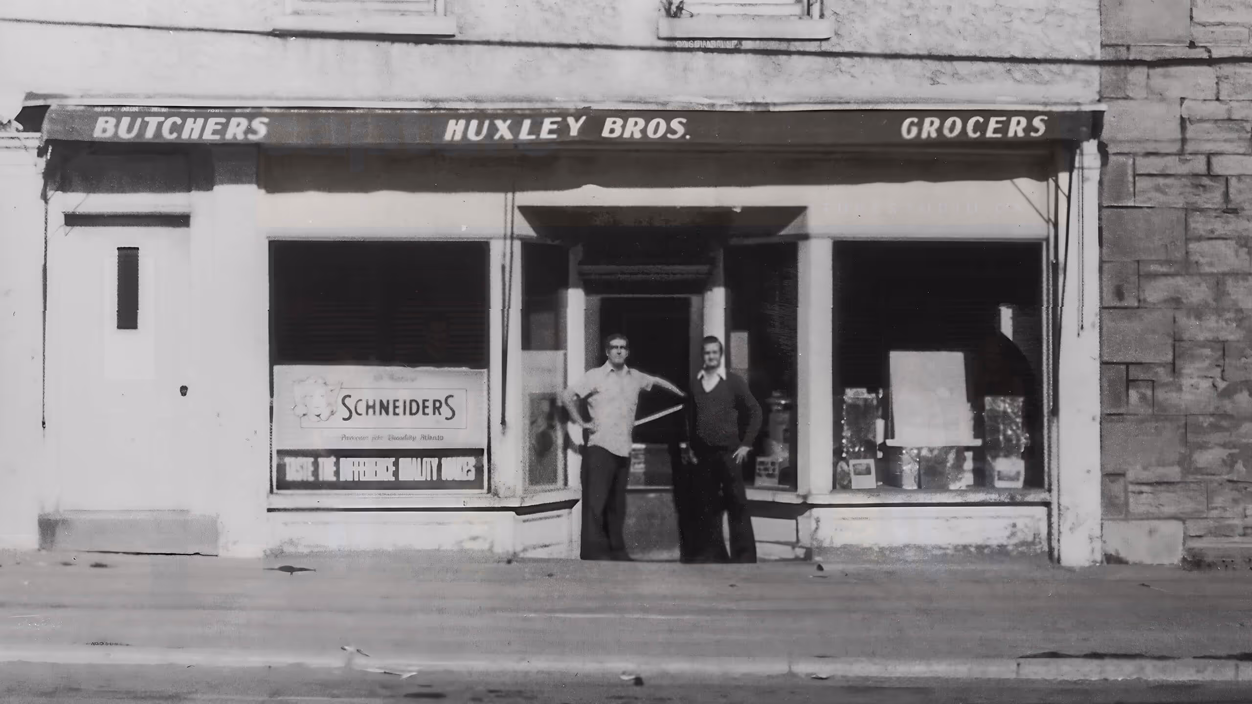 Black and white photo of two men standing in front of Huxley Bros. butcher and grocer shop.