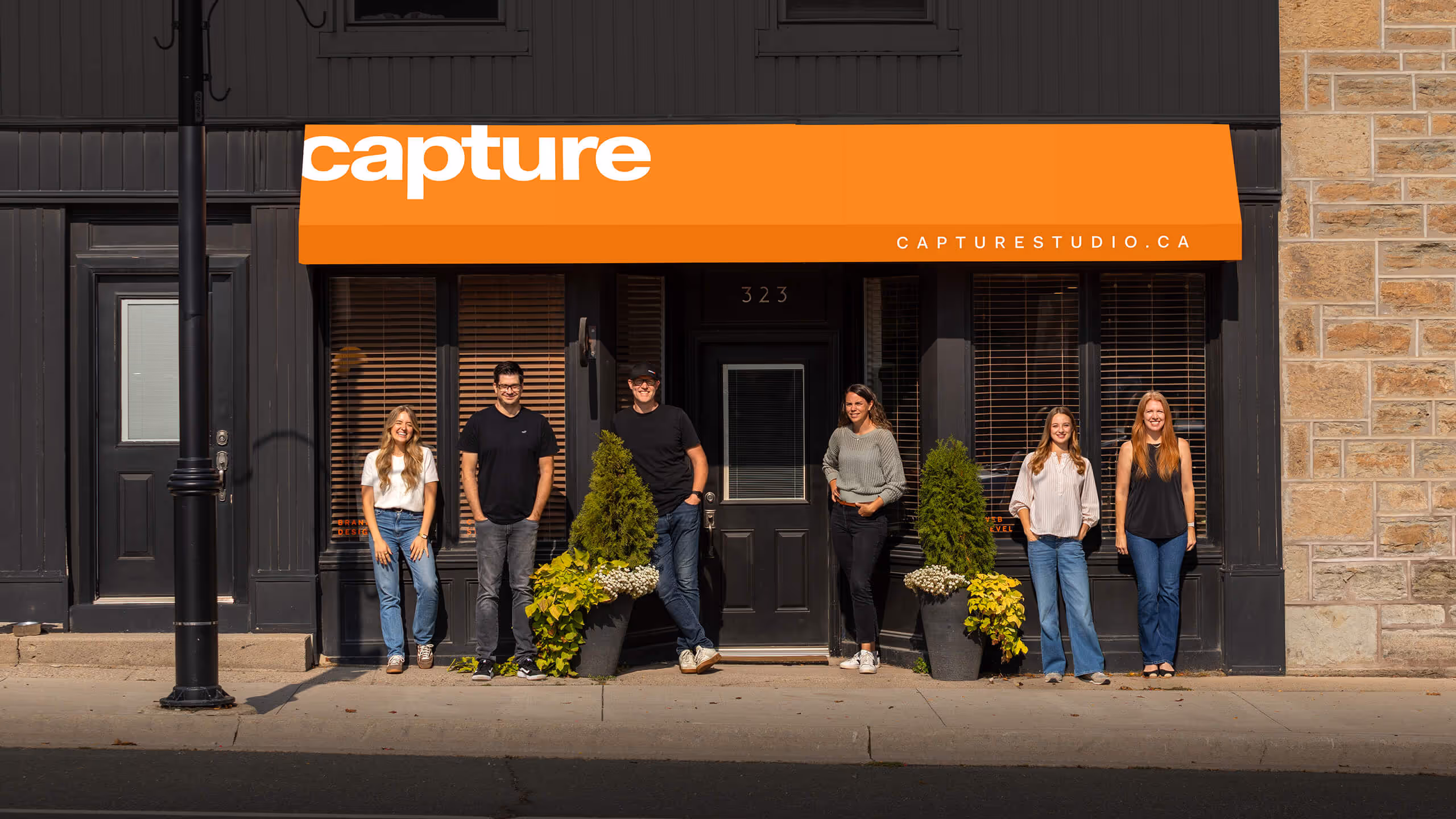 Six people standing in front of a black storefront with an orange awning that reads 'capture' and 'capturestudio.ca'.