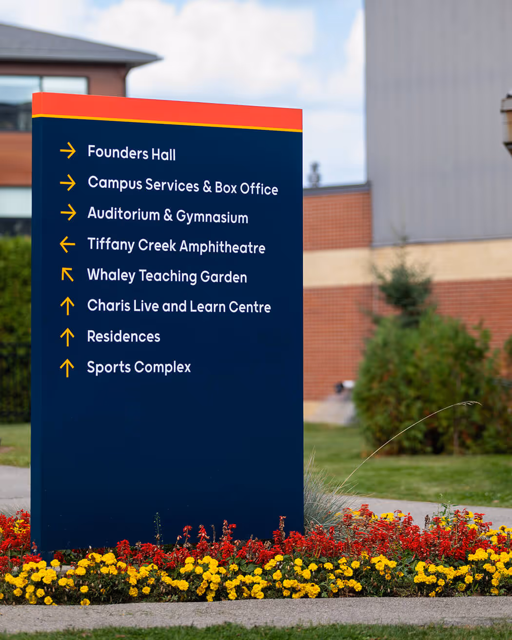 Campus directional sign listing locations including Founders Hall, Campus Services & Box Office, Auditorium & Gymnasium, Tiffany Creek Amphitheatre, Whaley Teaching Garden, Charis Live and Learn Centre, Residences, and Sports Complex, surrounded by red and yellow flowers.