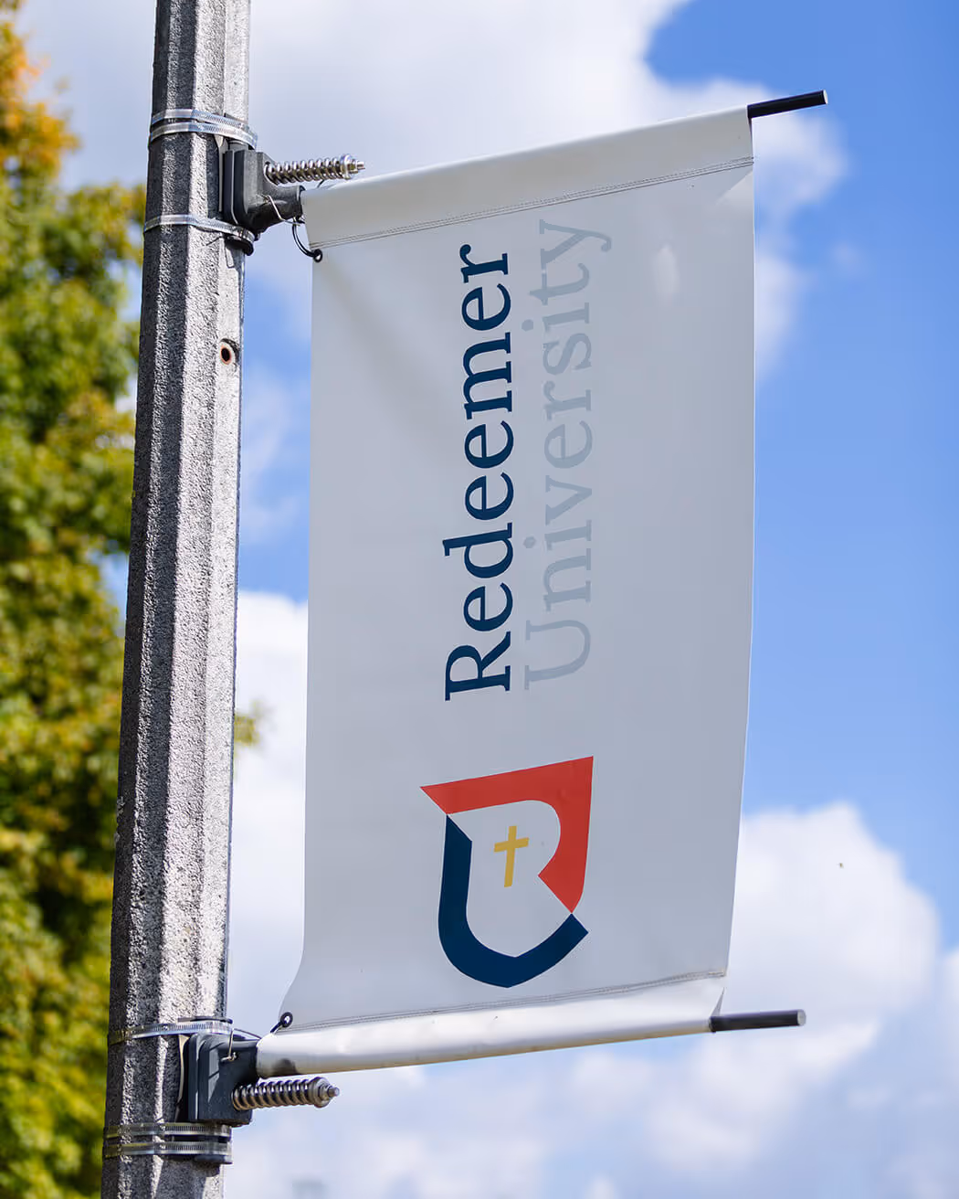 Flag banner on a pole displaying Redeemer University logo with a cross shield against a blue sky background.