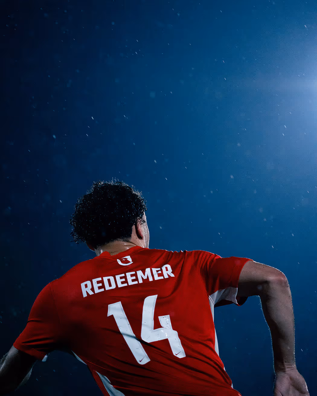 Soccer player wearing red jersey number 14 with the name Redeemer, seen from the back under night sky with rain.