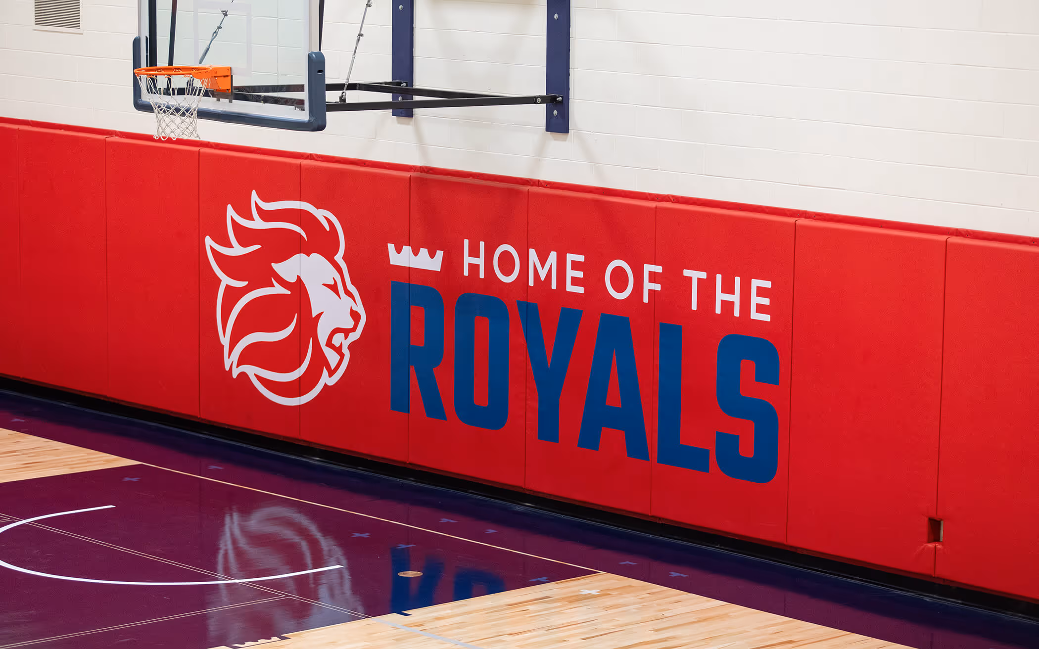Red padded gym wall with a lion logo and text reading 'Home of the Royals' beneath a basketball hoop.