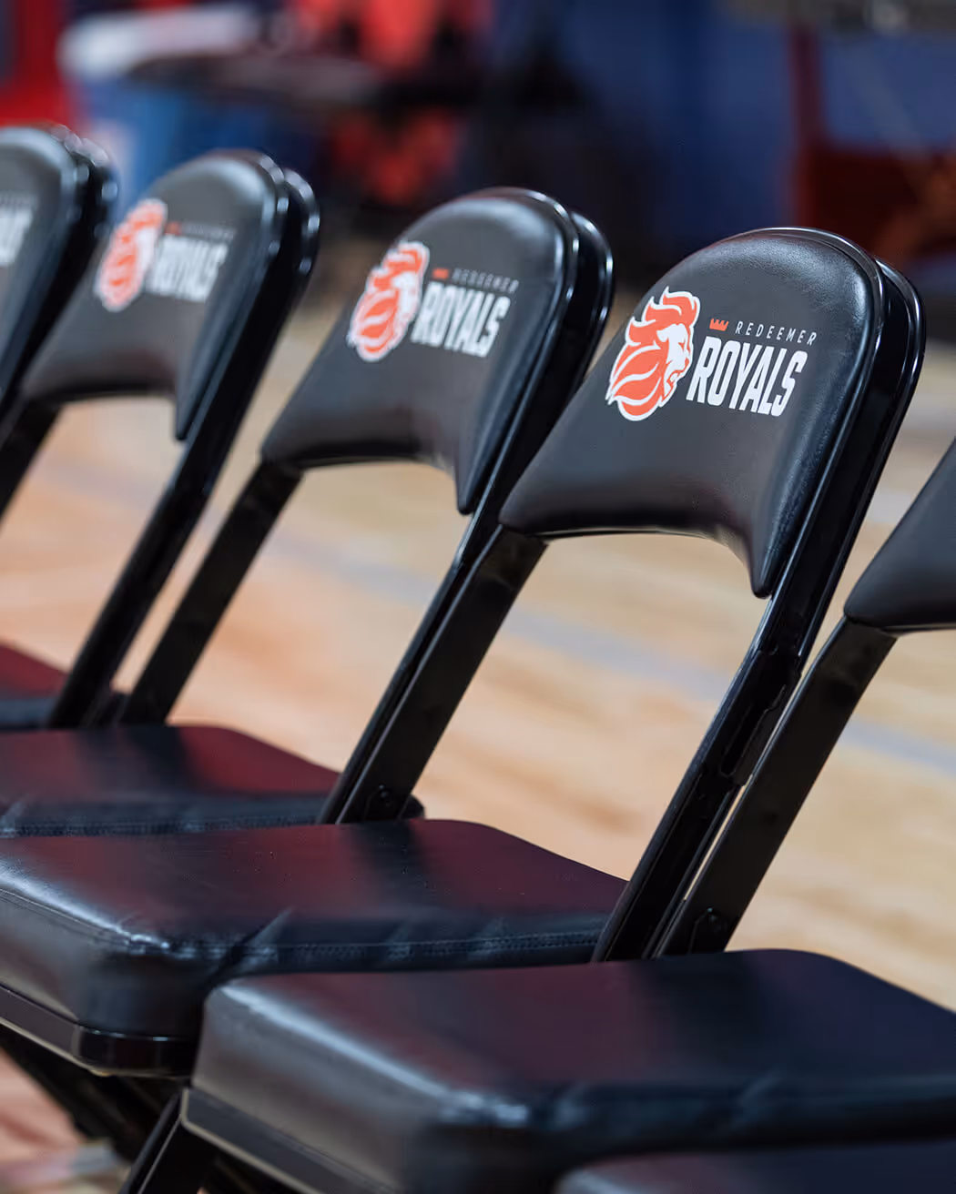 Row of black folding chairs with Redeemer Royals logo on the backrests arranged on a basketball court.