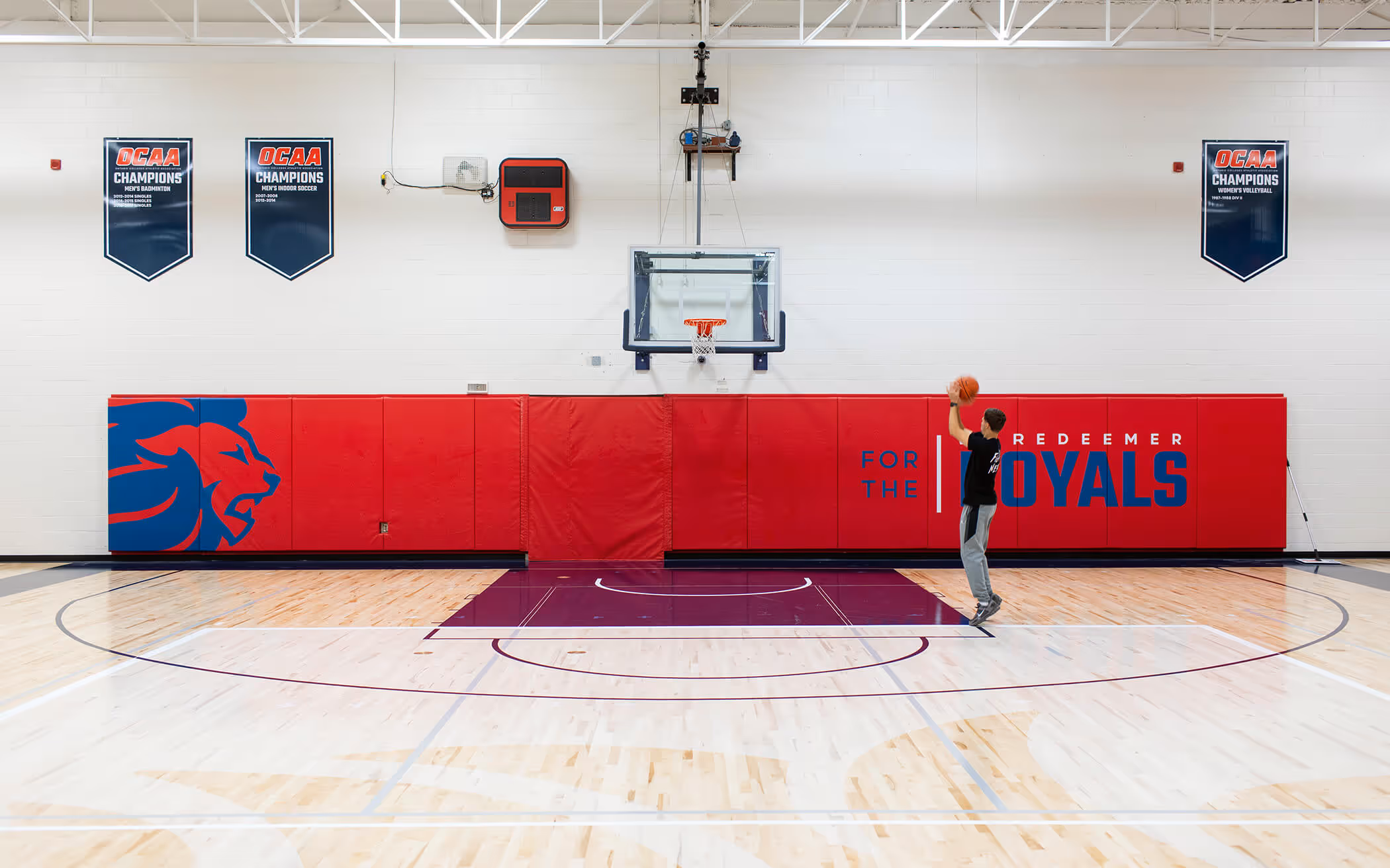 Young man in gym shooting basketball toward hoop on polished court with red padded wall and sports championship banners.