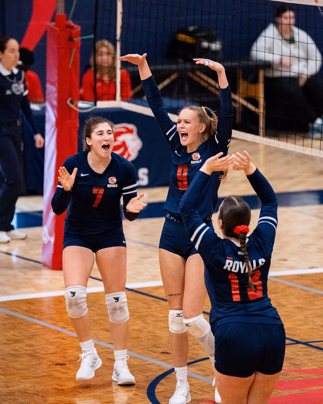 Three volleyball players in navy blue Royals uniforms celebrating on the court near the net.