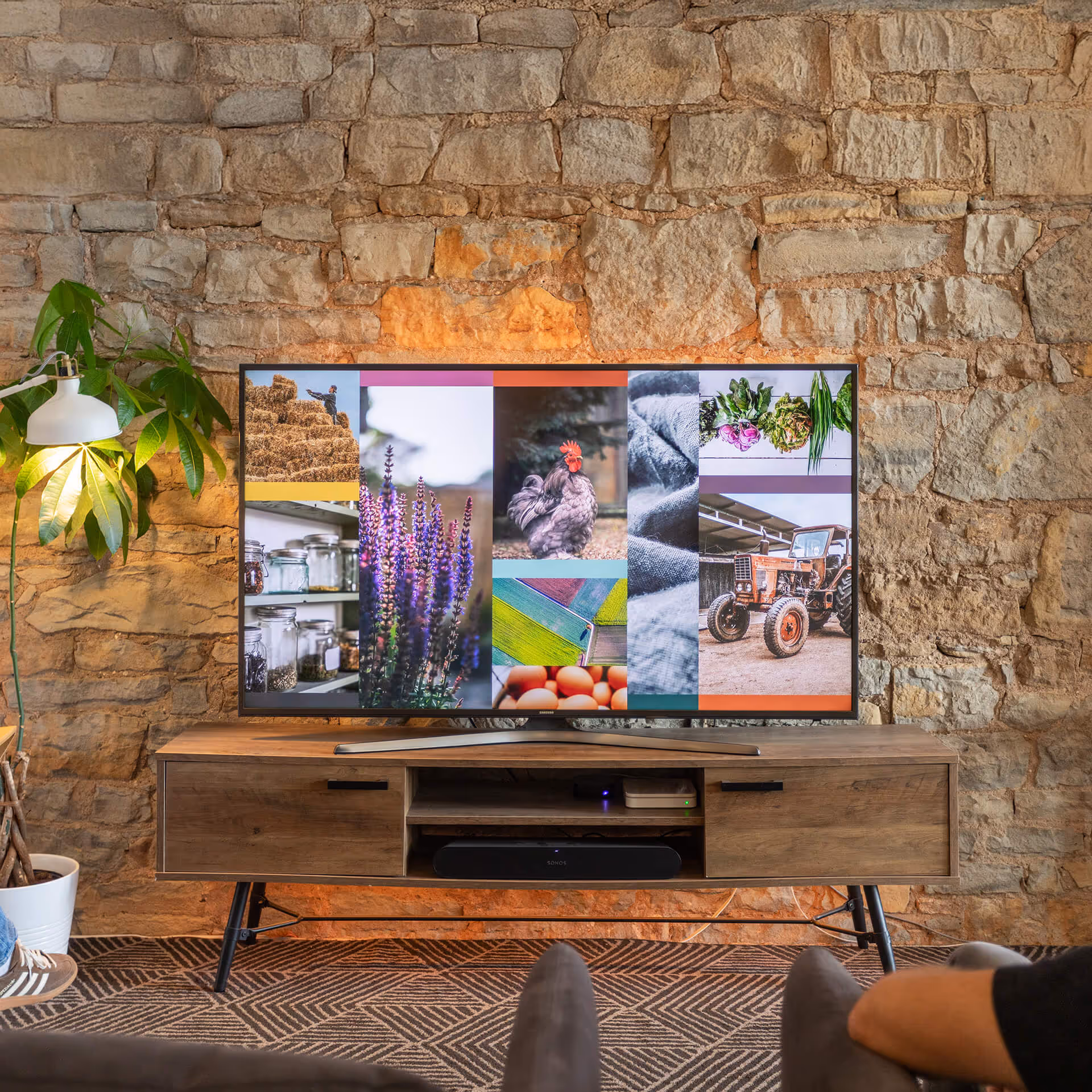 Modern living room setup with a flat-screen TV on a wooden stand, displaying a collage of farm-related images against a stone wall background.
