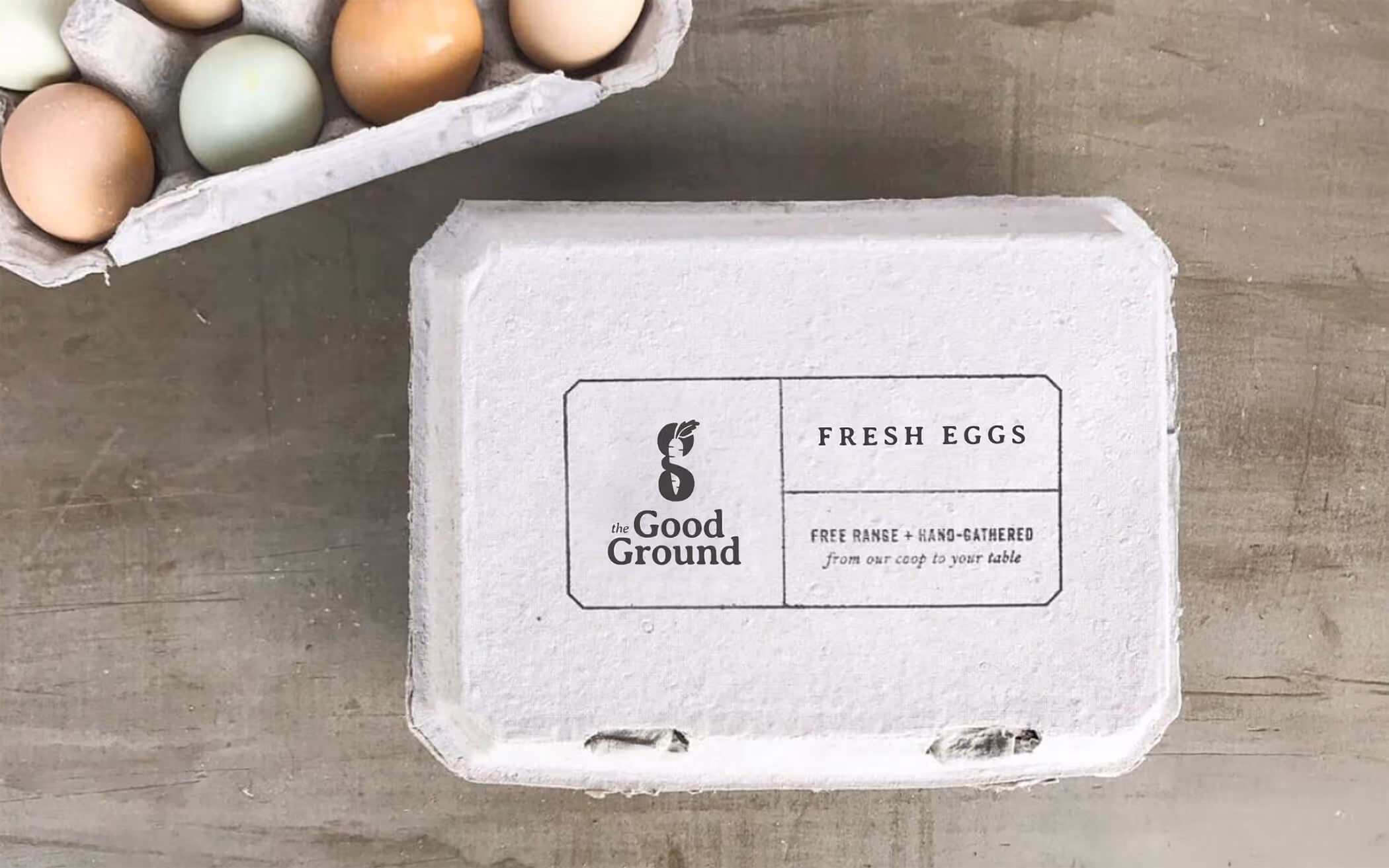 White egg carton with The Good Ground logo labeled 'Fresh Eggs' and 'Free Range + Hand-Gathered from our coop to your table' next to a carton of multicolored eggs.